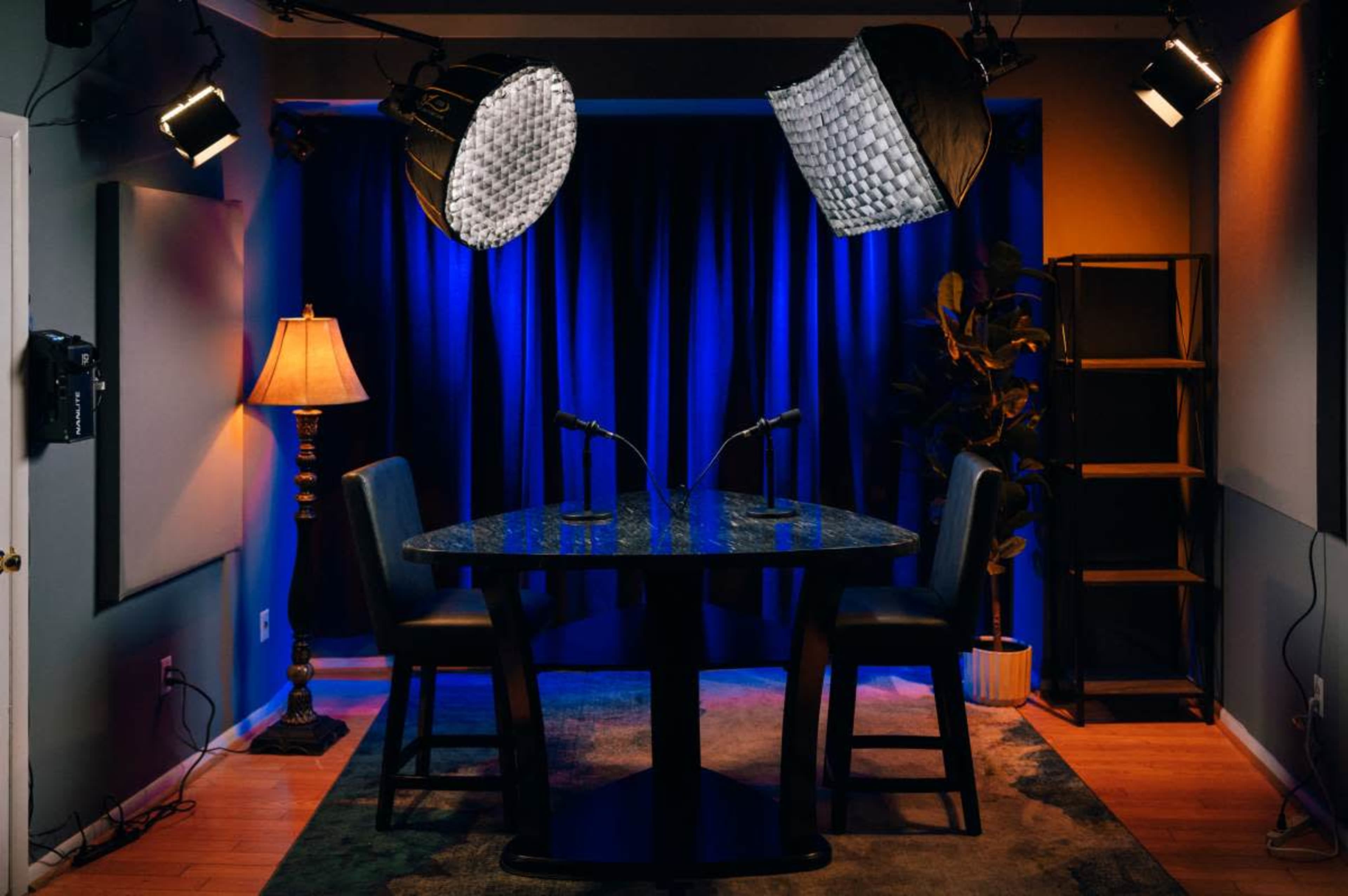 A two-person interview setup features a round table with microphones, a well-lit backdrop, and a lamp in a dimly lit room.