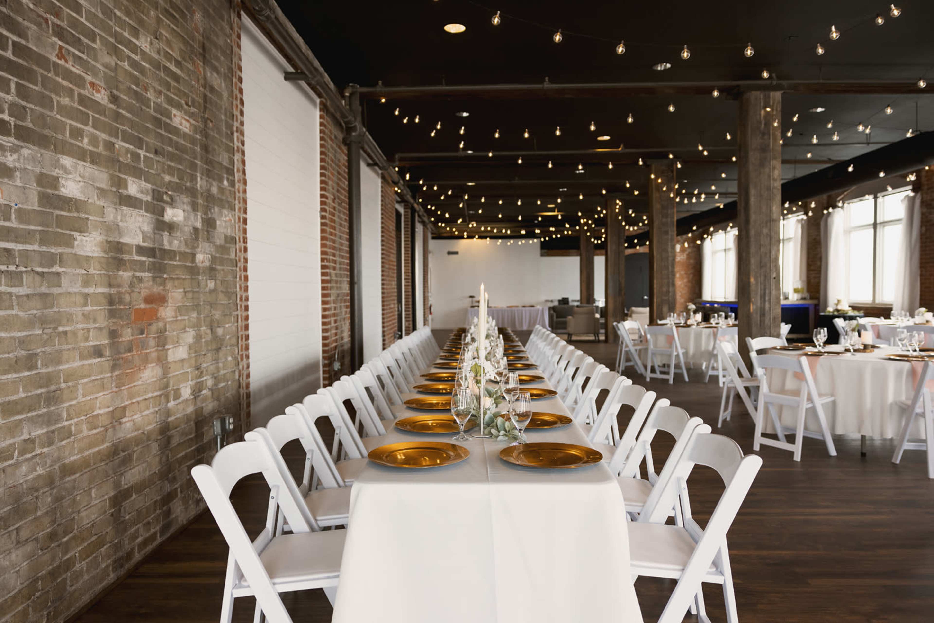 A long banquet table is elegantly arranged with white tablecloths and gold-rimmed plates, set in a well-lit industrial space featuring exposed brick walls and string lights overhead.