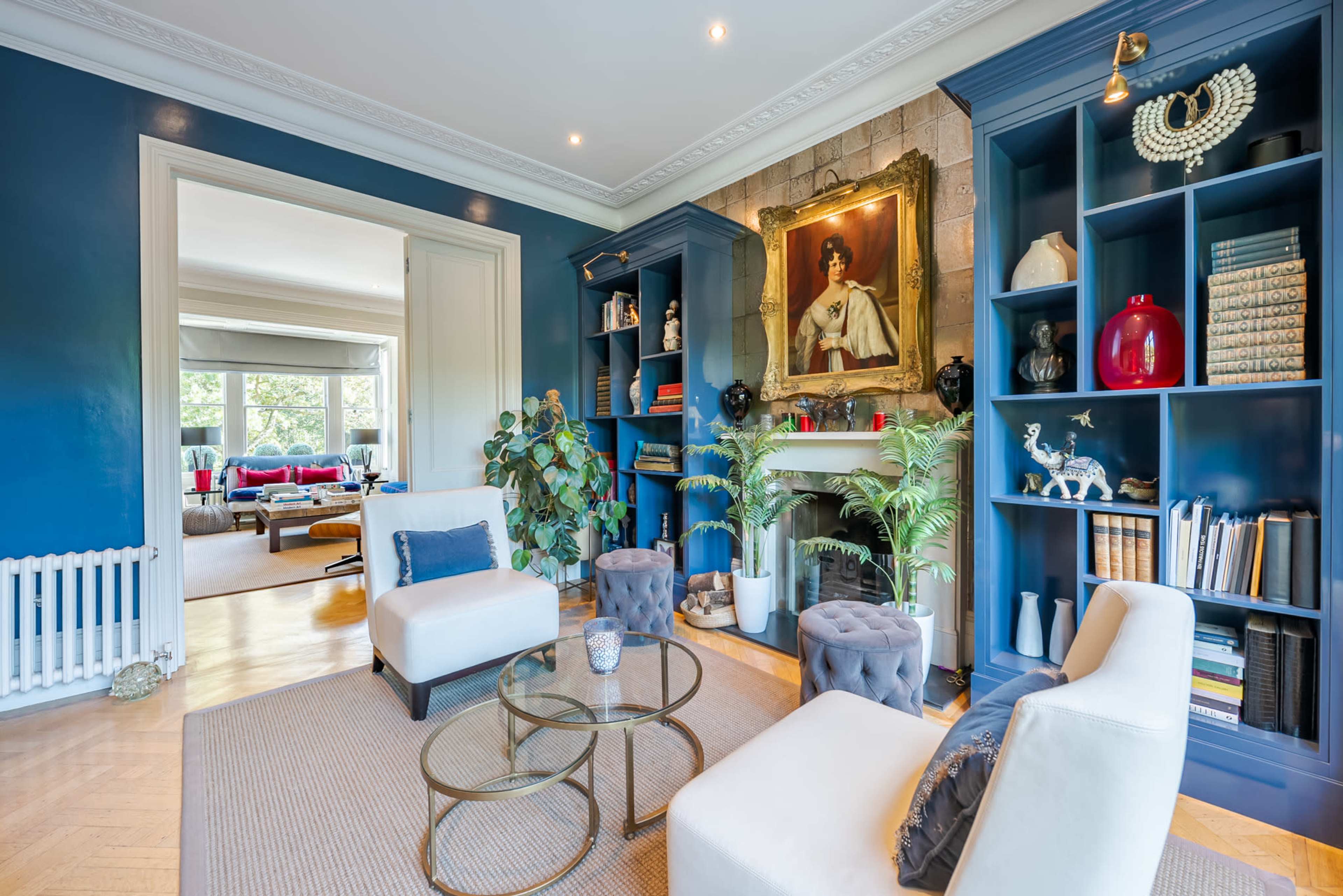 The image shows a living room with blue walls, two white chairs around a glass coffee table, and a fireplace adorned with plants and decor.