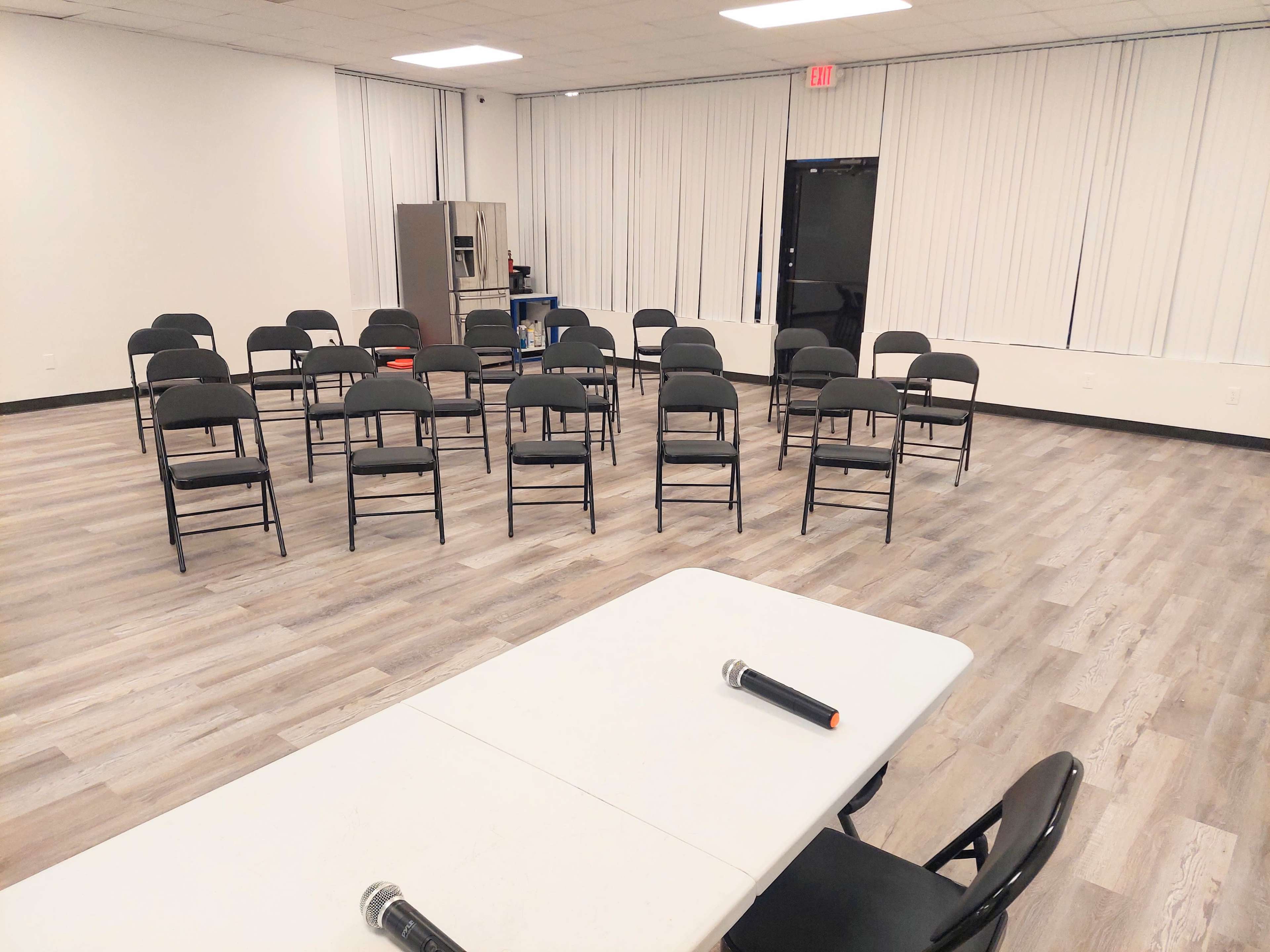 A room with rows of black chairs arranged in a circular formation, accompanied by a long white table at the front with two microphones on it.