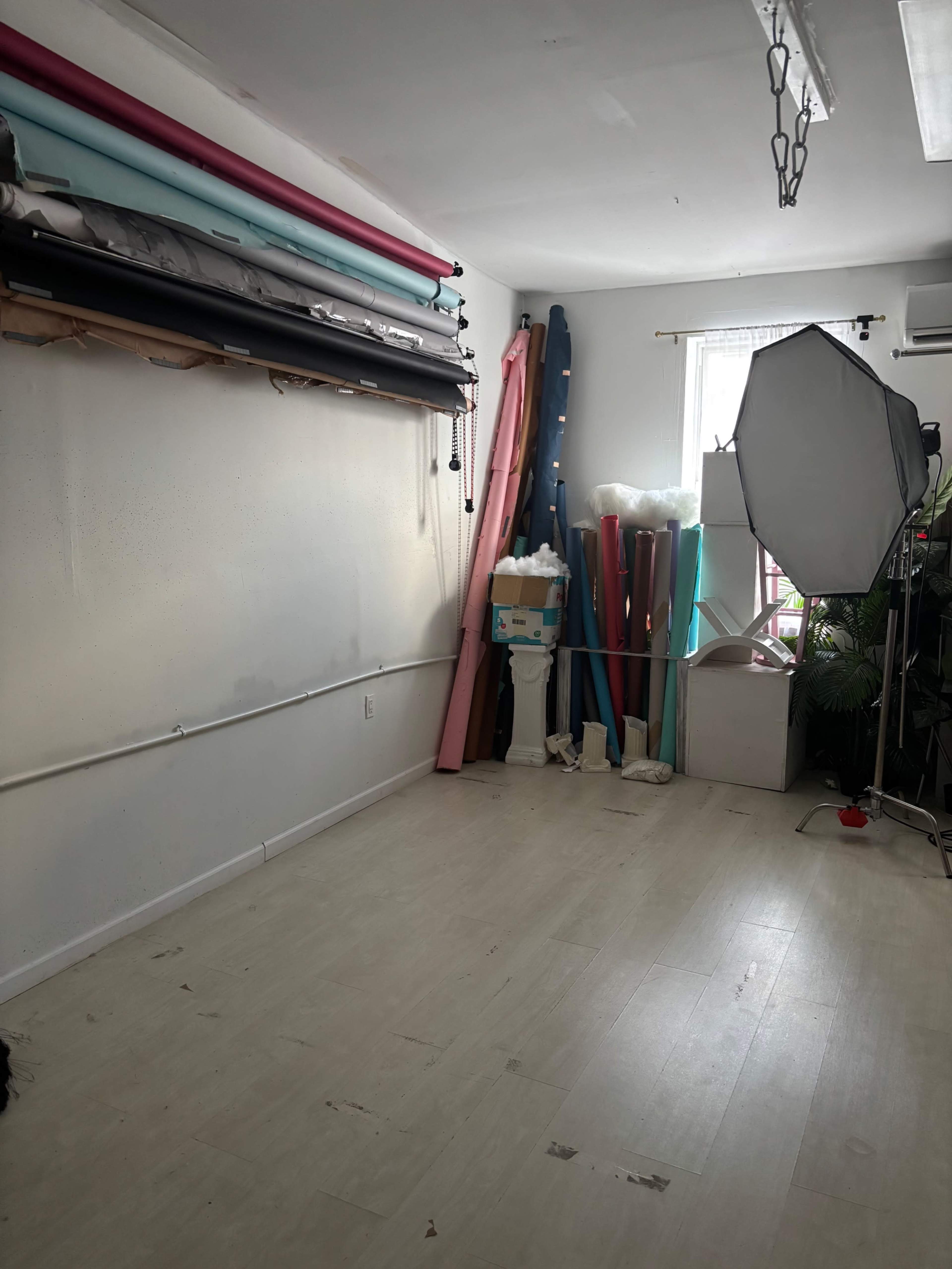 The image shows a photography studio with a plain wall, colorful backdrops hanging on rails, and a lighting setup in the corner.