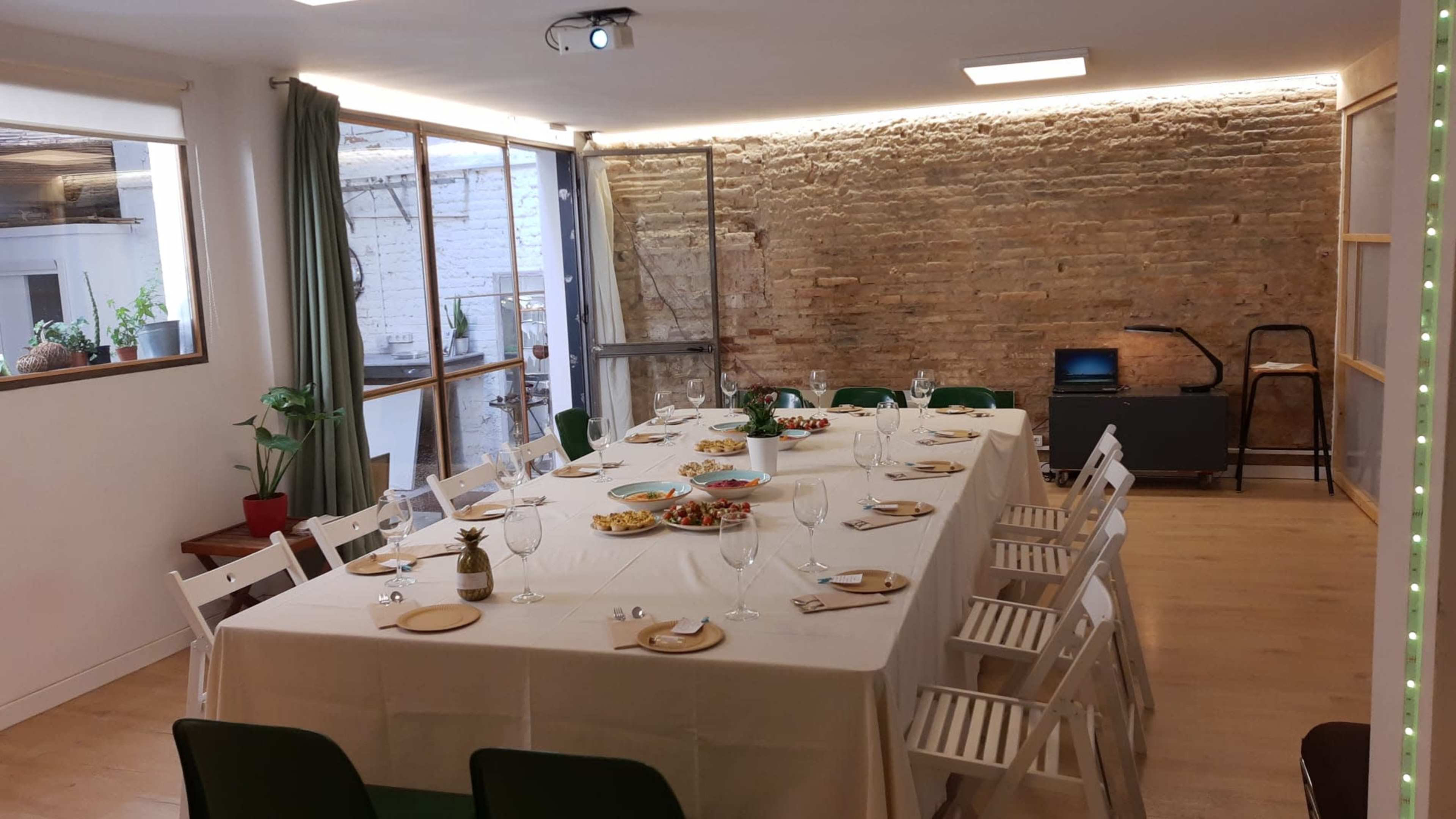 A long dining table set for a meal is positioned in a well-lit room with a brick wall and large windows.