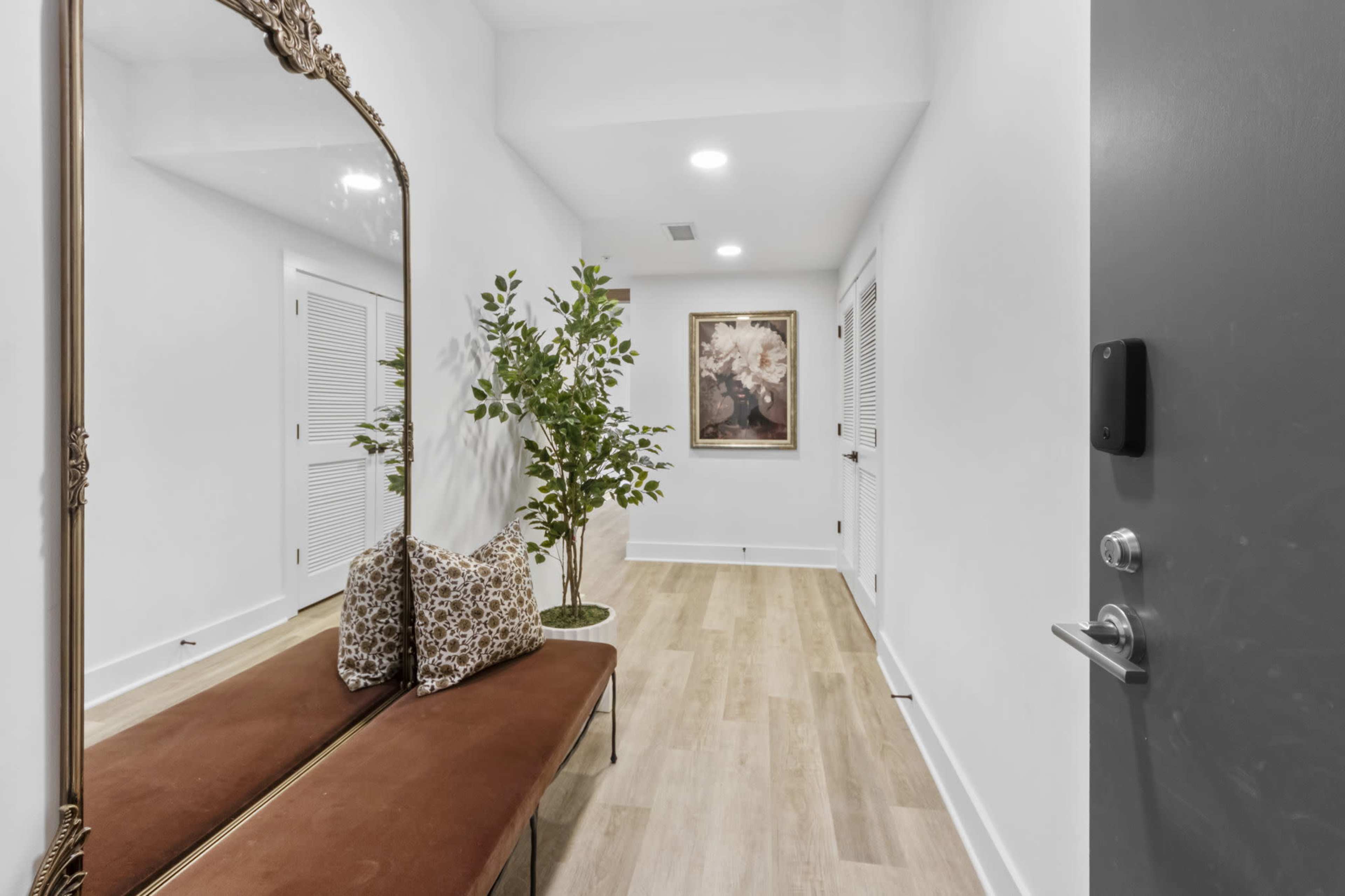 The image shows a spacious hallway featuring a large decorative mirror, a brown bench with patterned pillows, a potted plant, and a framed artwork on the wall.