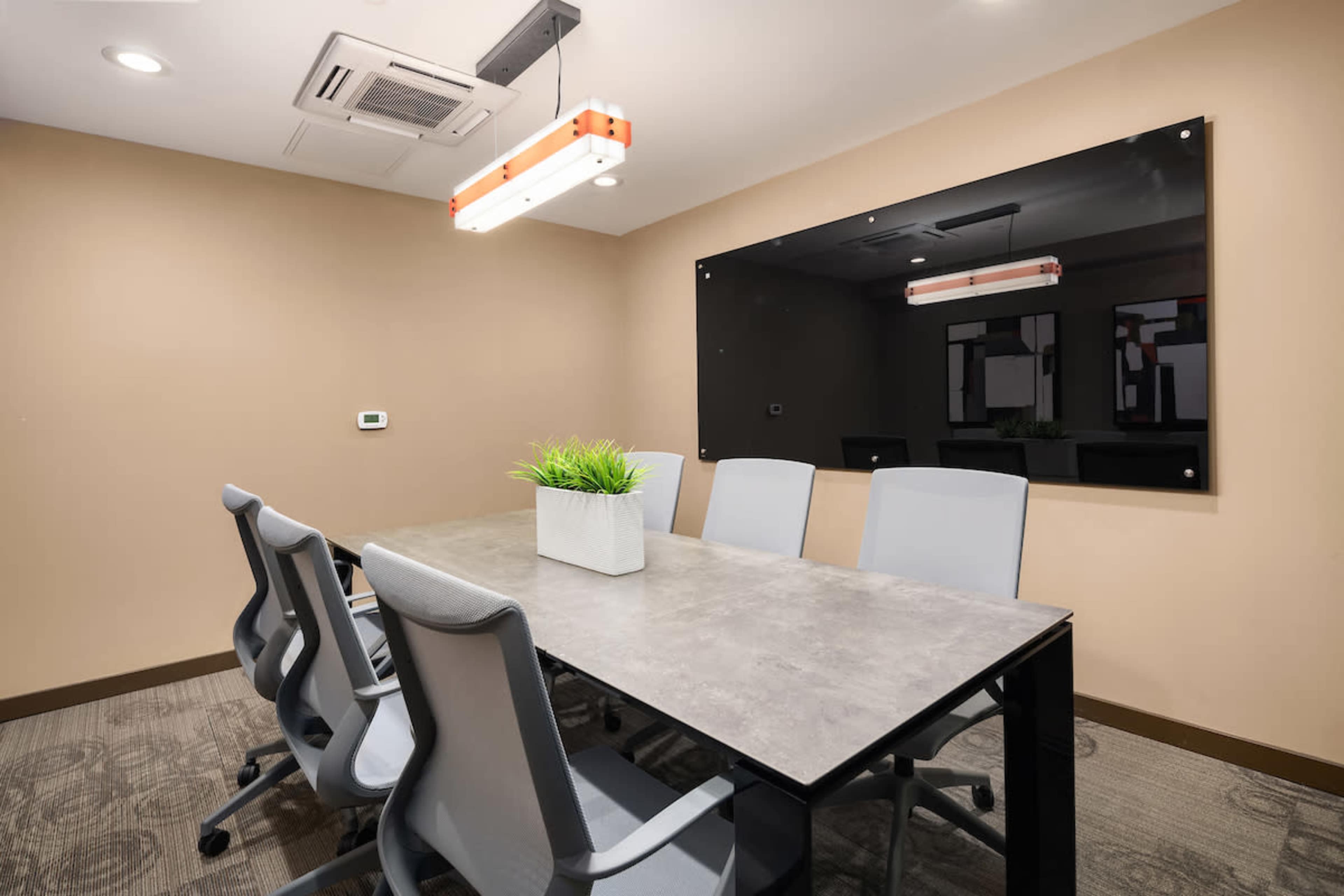 The image shows a modern conference room with a rectangular table surrounded by six gray chairs and a wall-mounted black board.