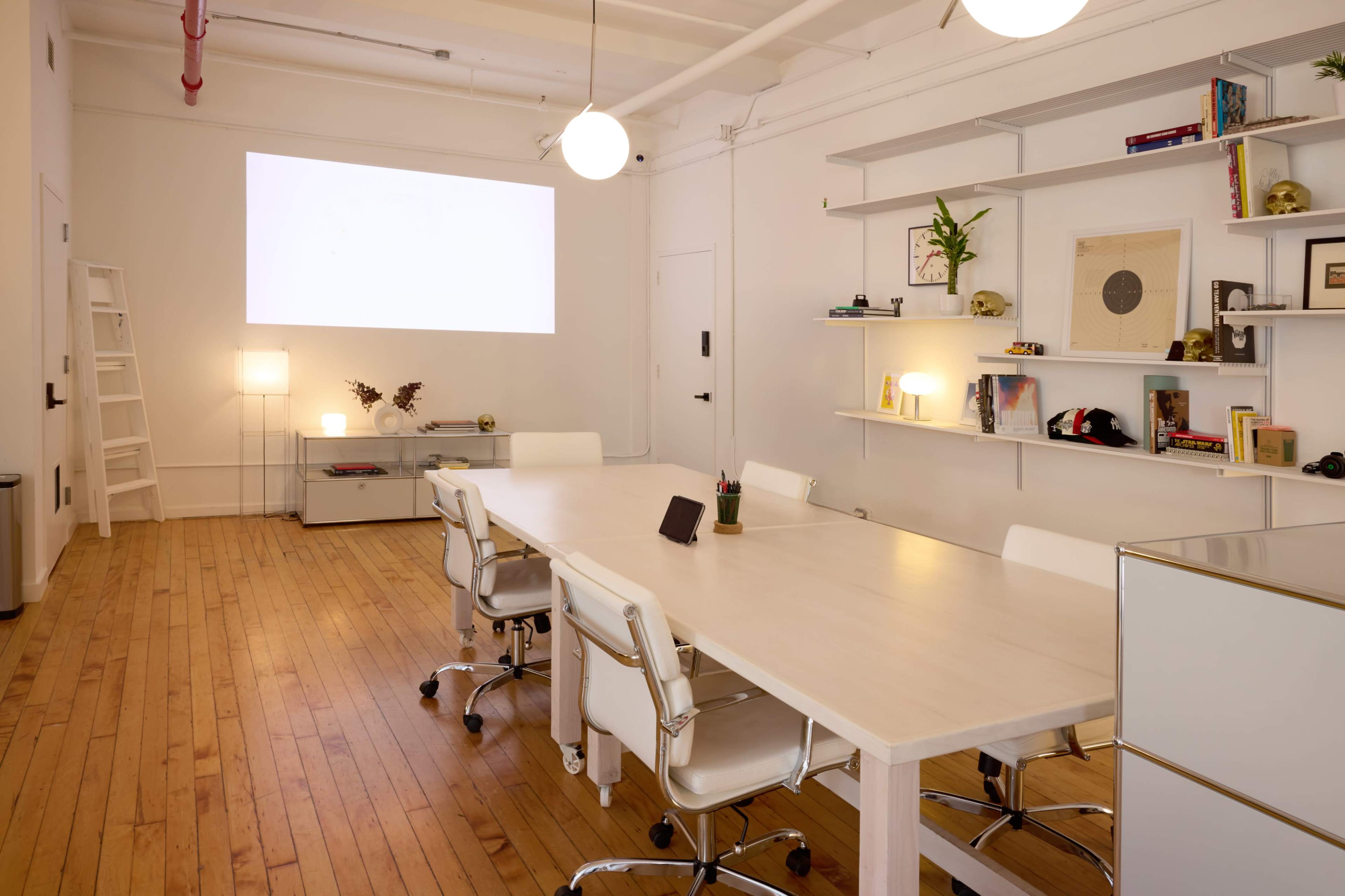 A modern conference room features a long table surrounded by white chairs, with a projector screen on the wall and shelves displaying decorative items.