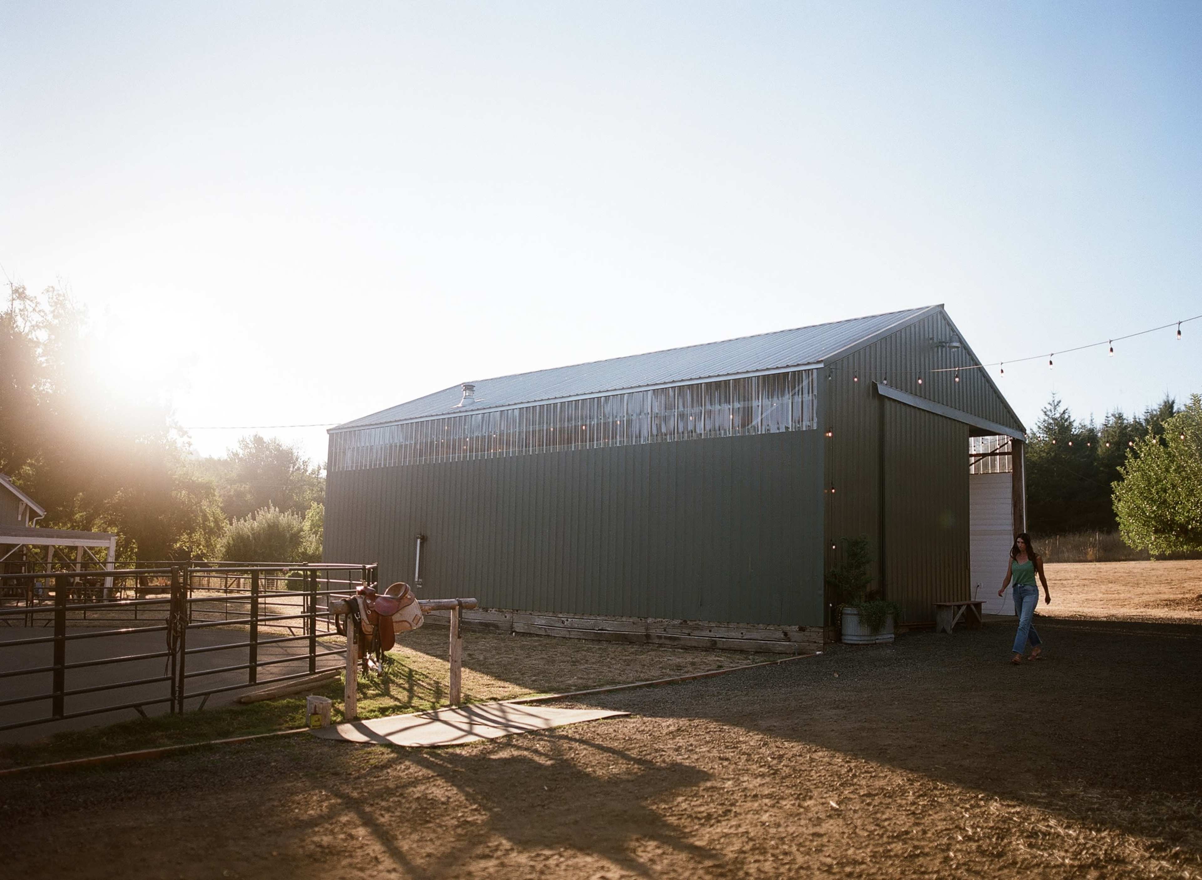 A metal barn stands in a rural landscape under a clear blue sky, with a person walking toward it.