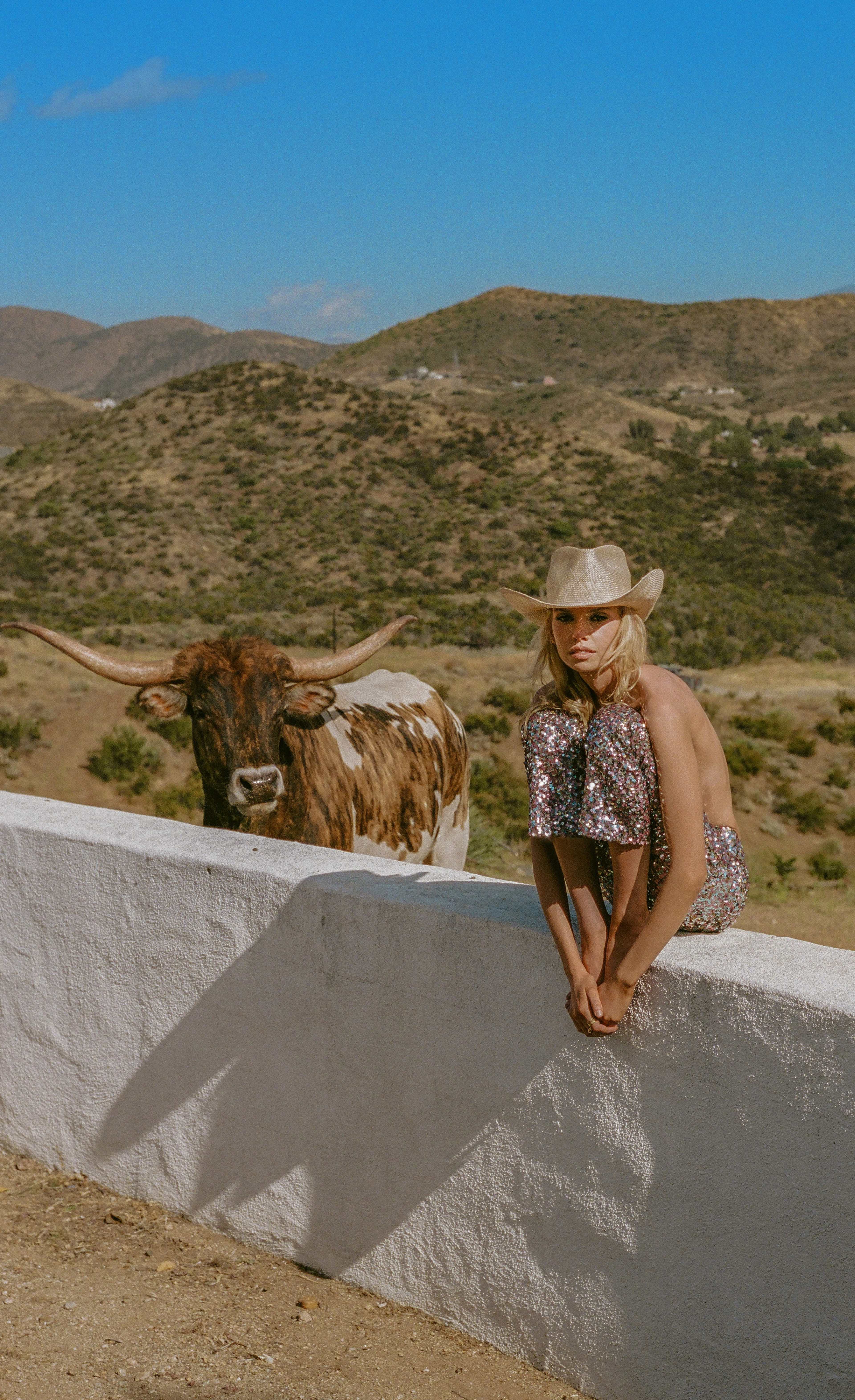 A woman in a sequined outfit sits on a white wall next to a longhorn cow with mountainous terrain in the background.