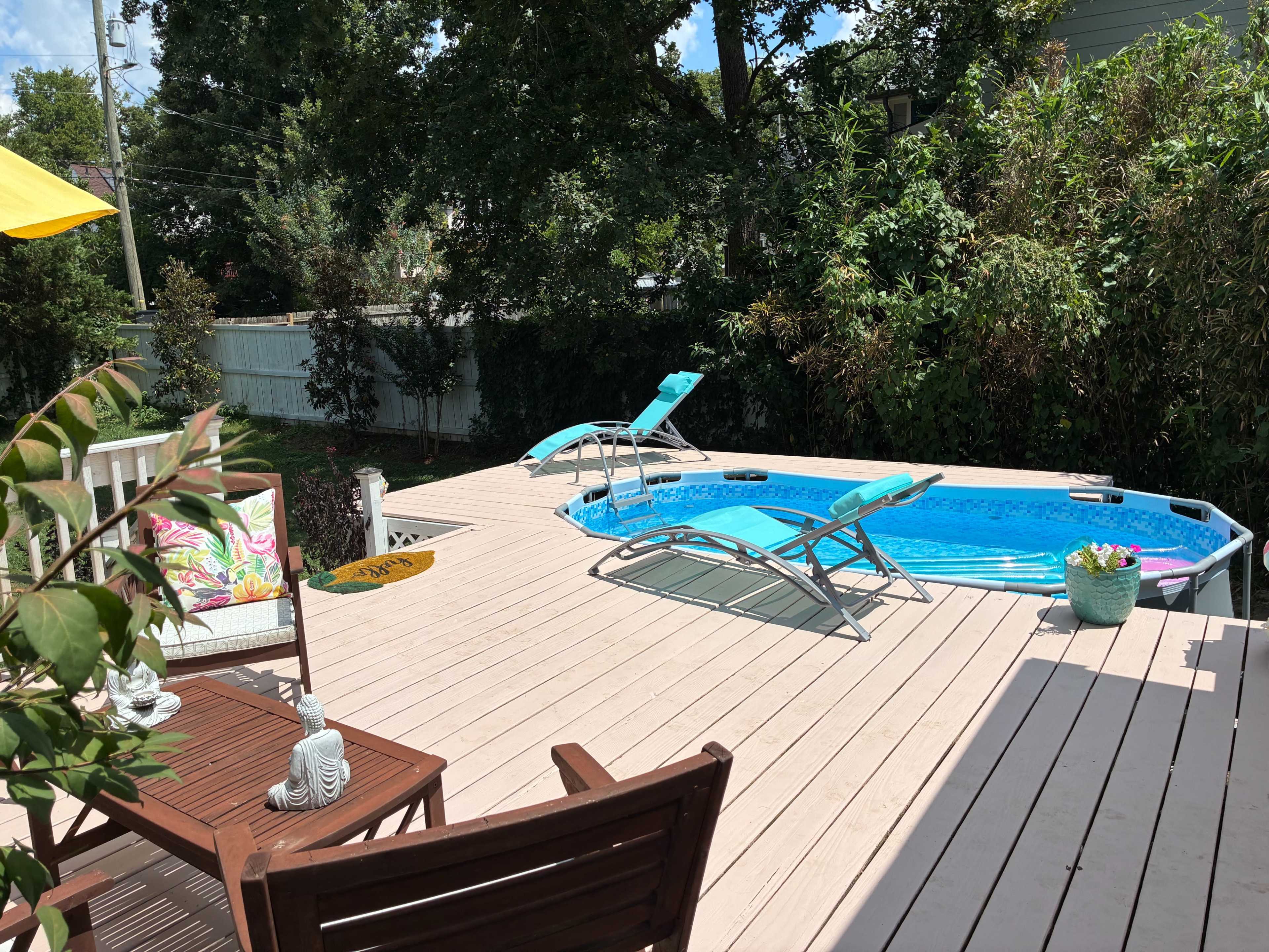 The image shows a backyard deck with a small swimming pool, two lounge chairs, and a seating area surrounded by greenery.
