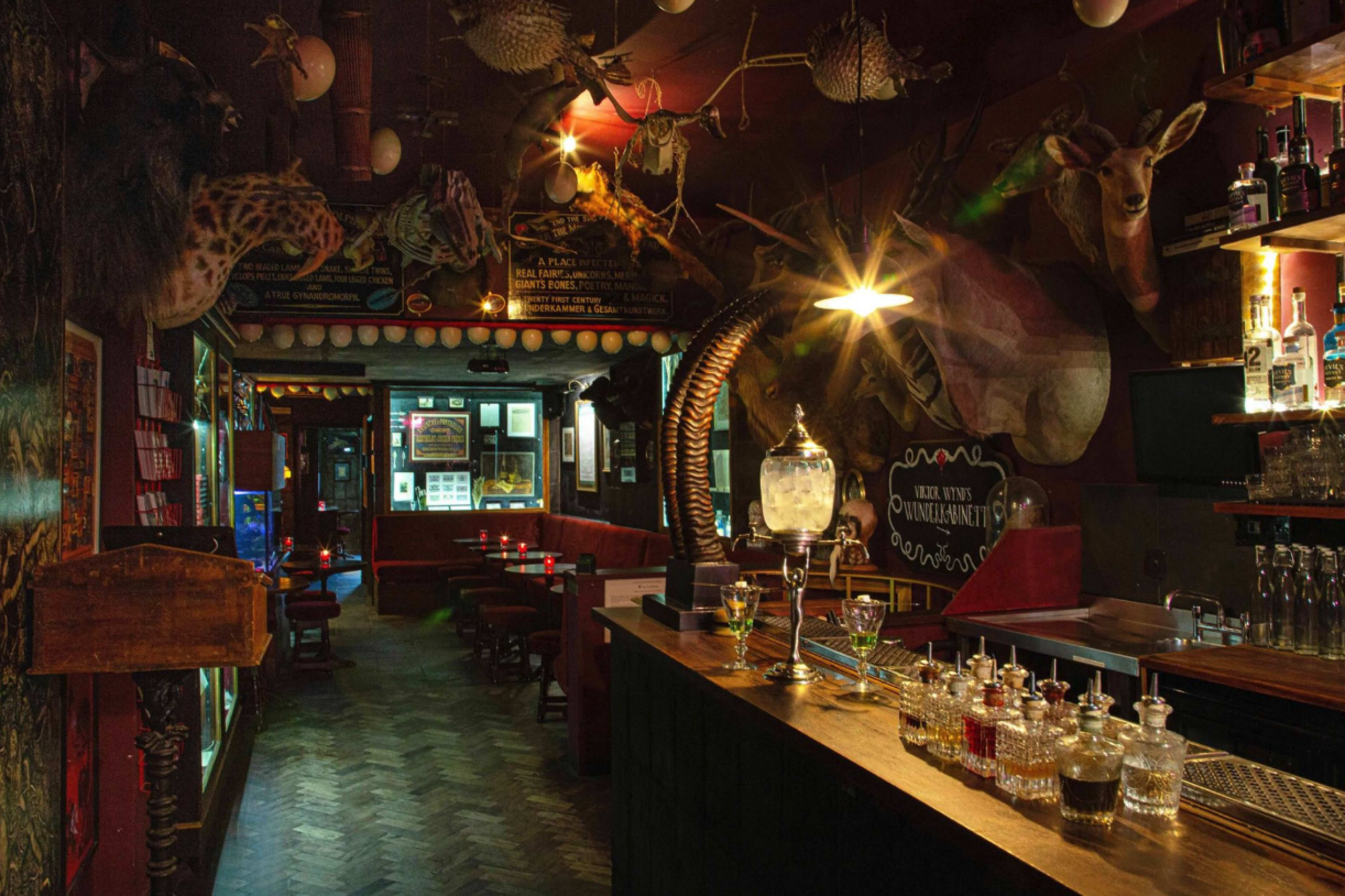 The image shows a dimly lit bar decorated with mounted animal heads and a long wooden counter lined with glass bottles and cocktail ingredients.
