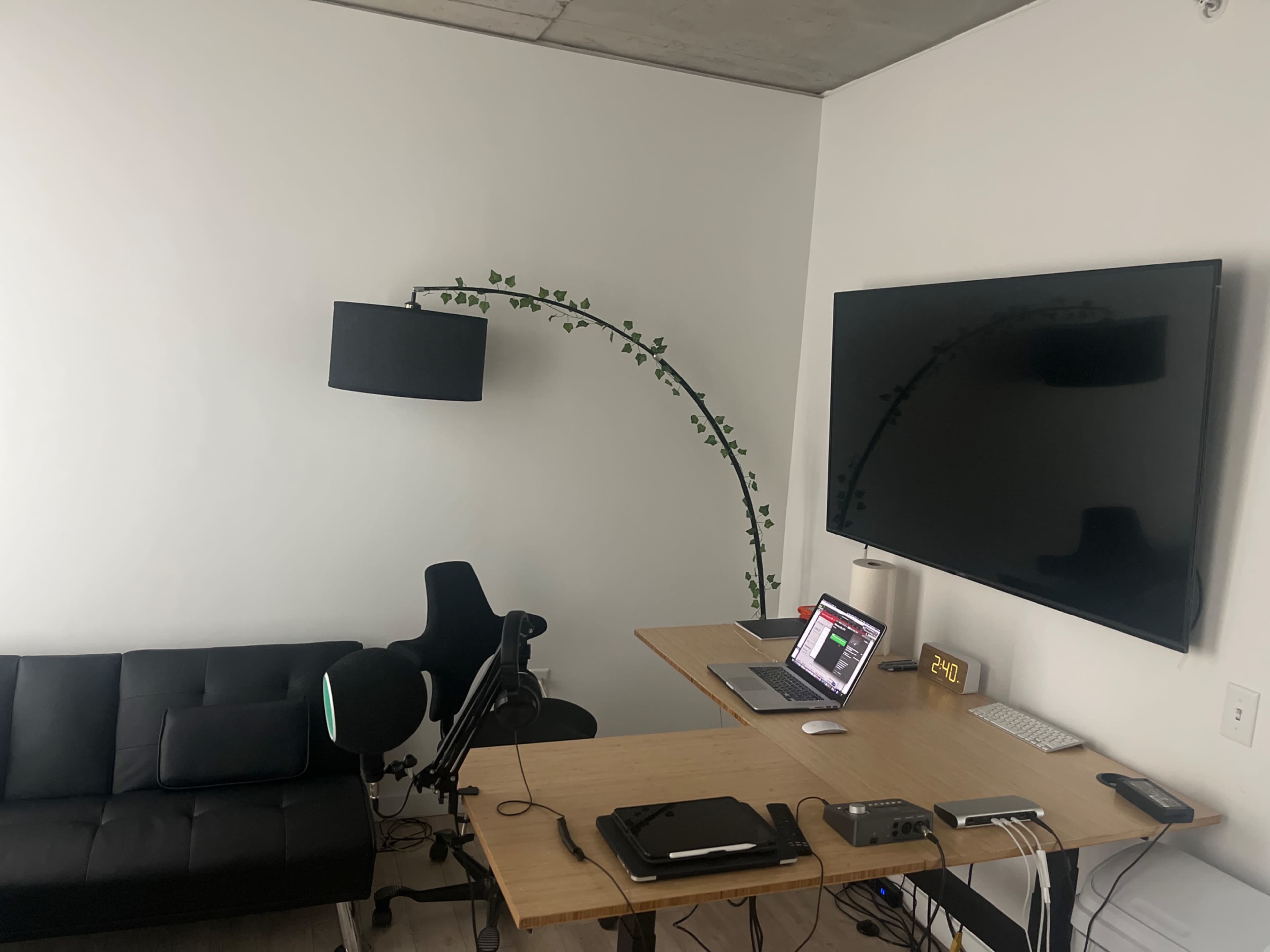 A minimalist home office with a large TV mounted on the wall, a wooden desk with a laptop and audio equipment, a black couch, and a lamp with a plant extending towards the wall.