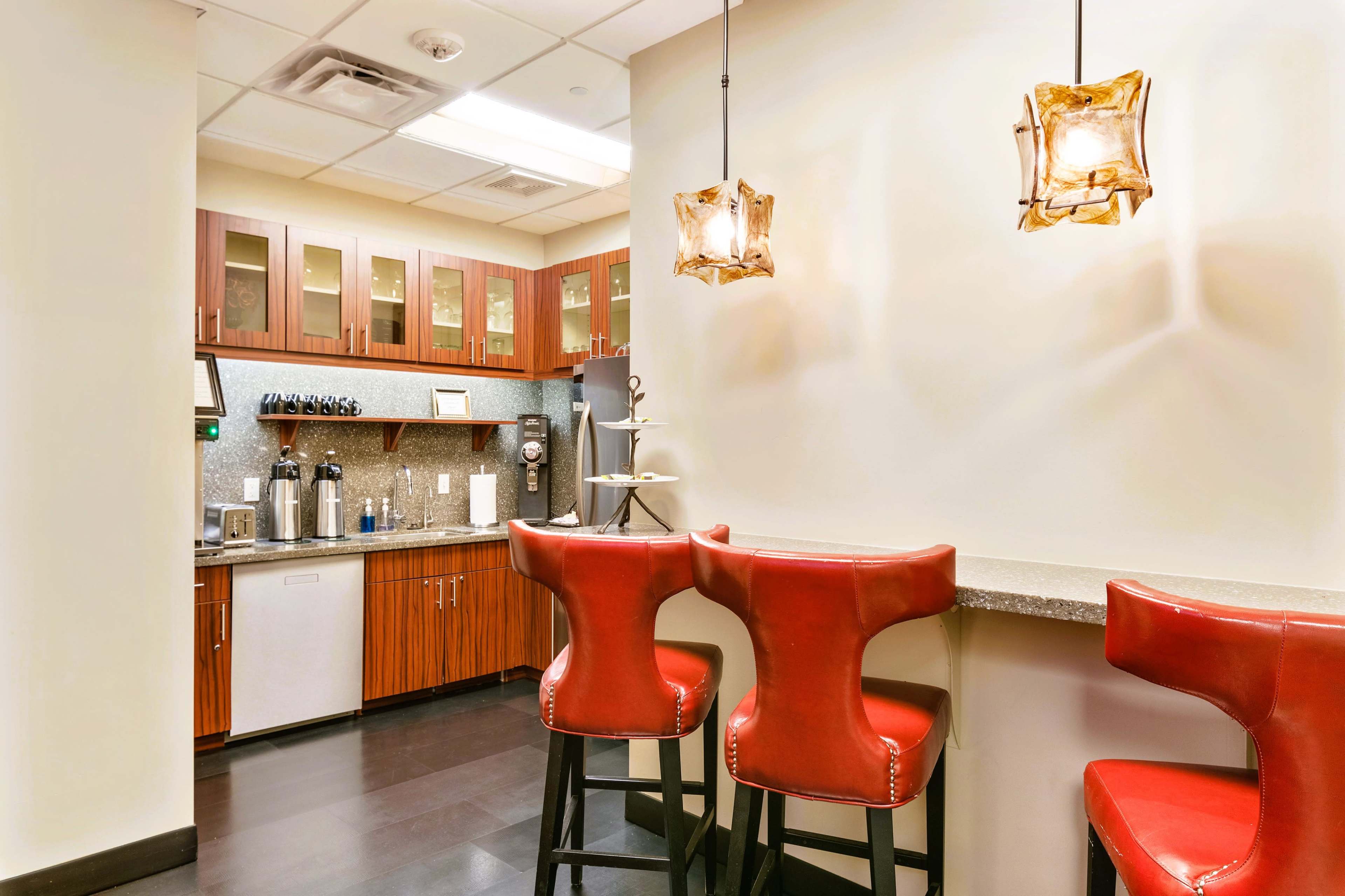 The image shows a modern kitchen area with wooden cabinets, a gray countertop, and red bar stools along a counter.