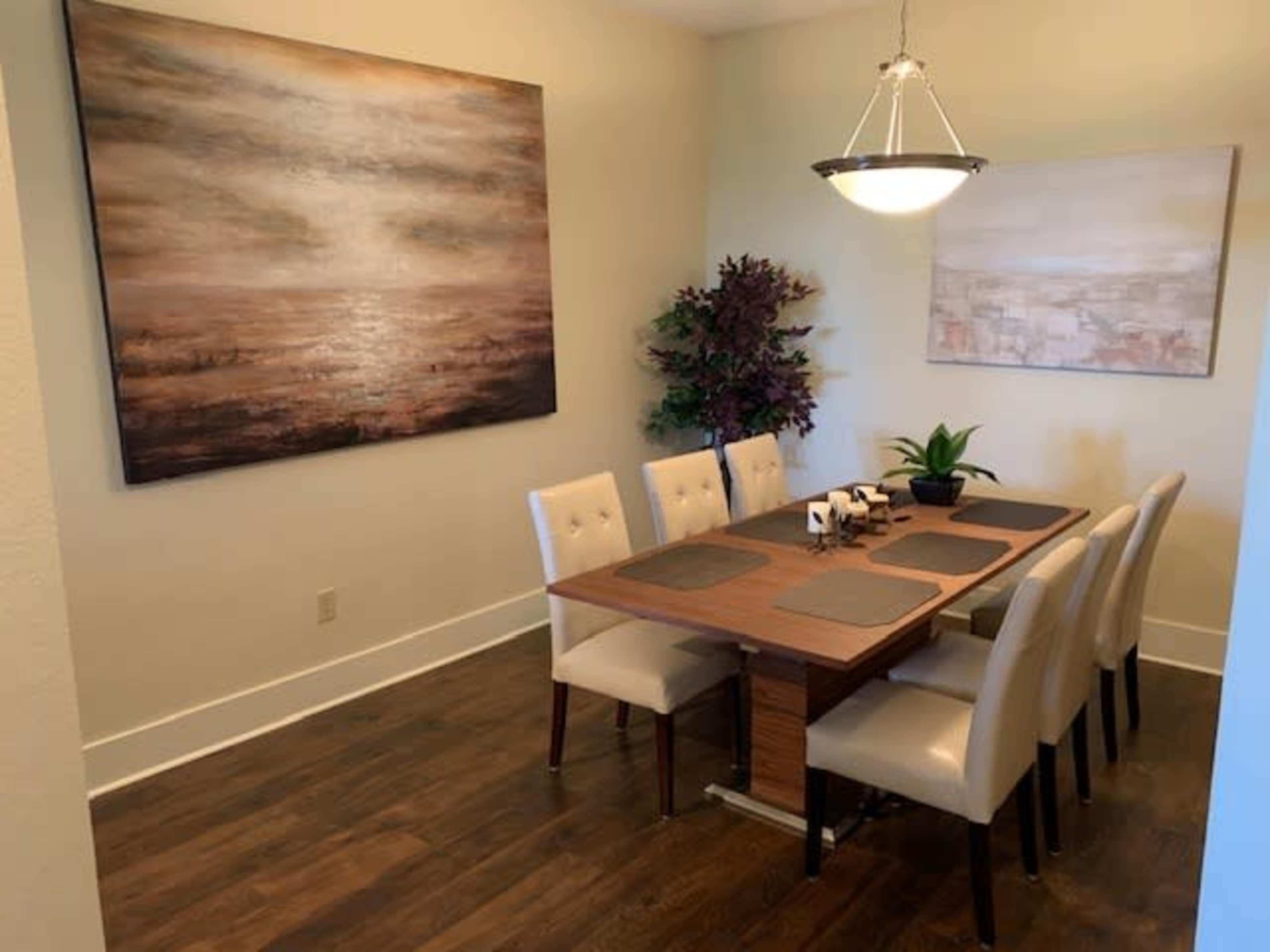 The image shows a dining room with a long wooden table surrounded by six upholstered chairs, decorated with artwork on the walls and a plant in the center.