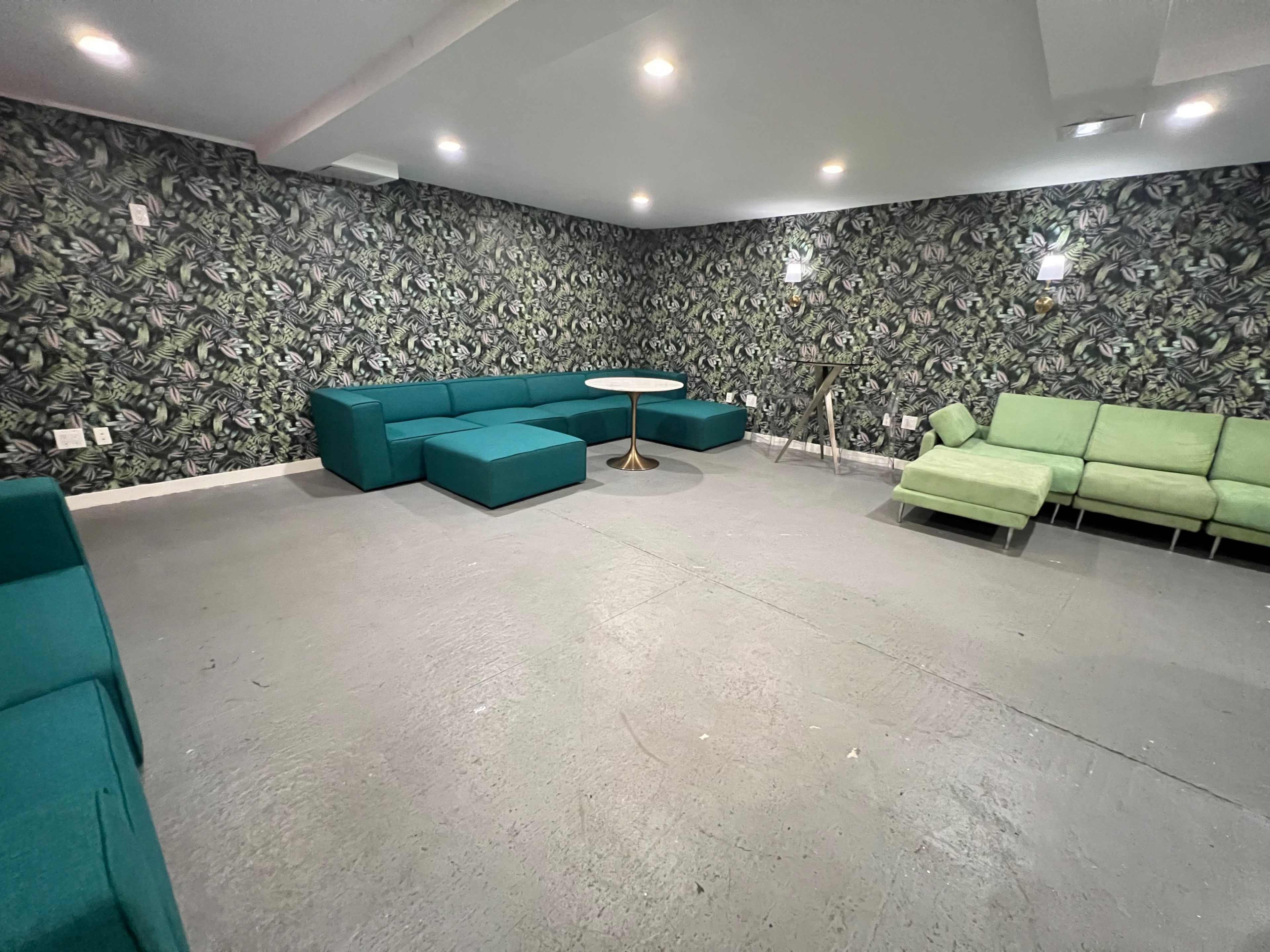 A spacious room with a patterned green wallpaper, featuring several teal sofas, a round table, and minimalist lighting.