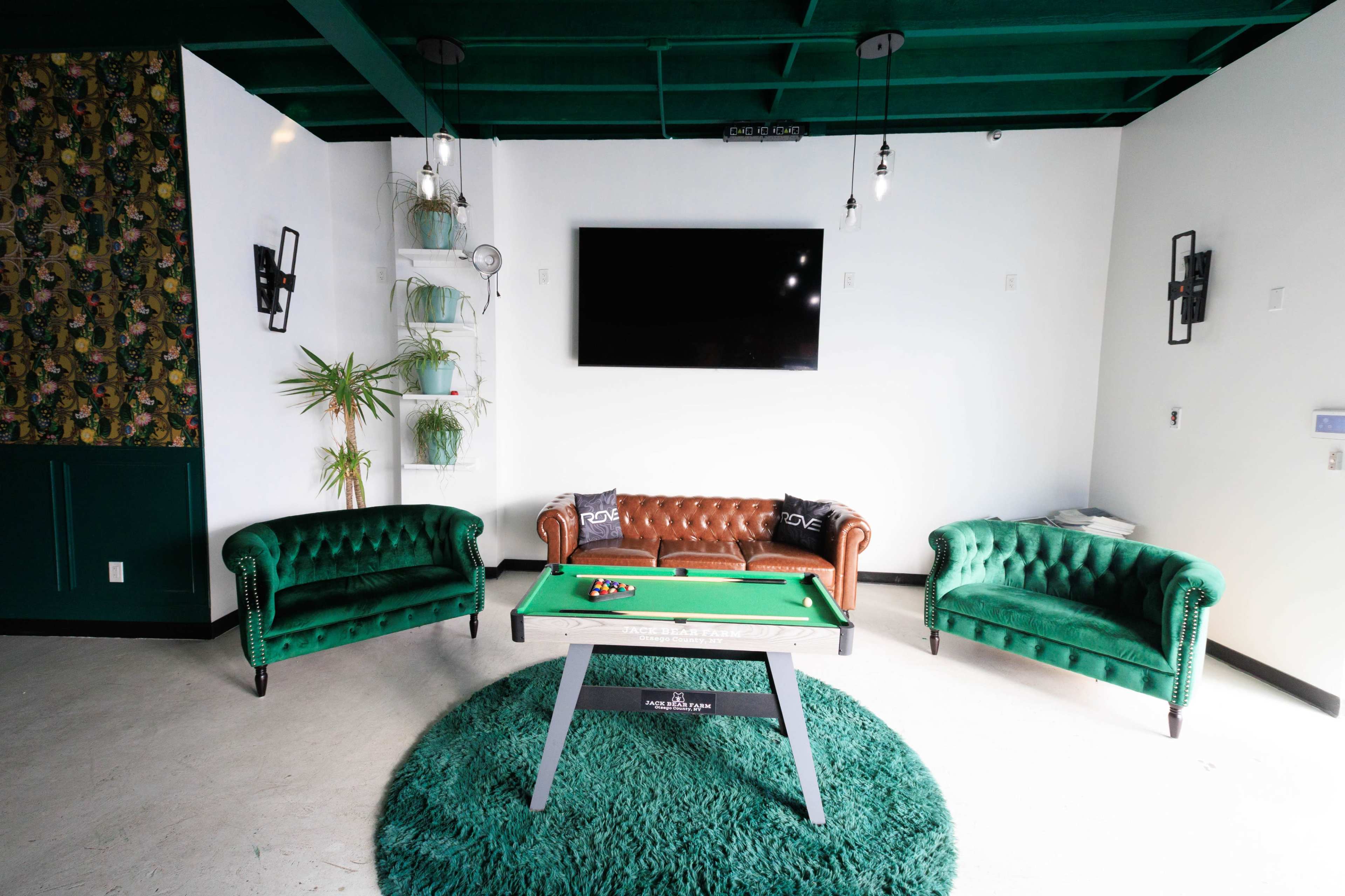 The image shows a stylish lounge area with green tufted sofas, a pool table in the center, and a large wall-mounted television.