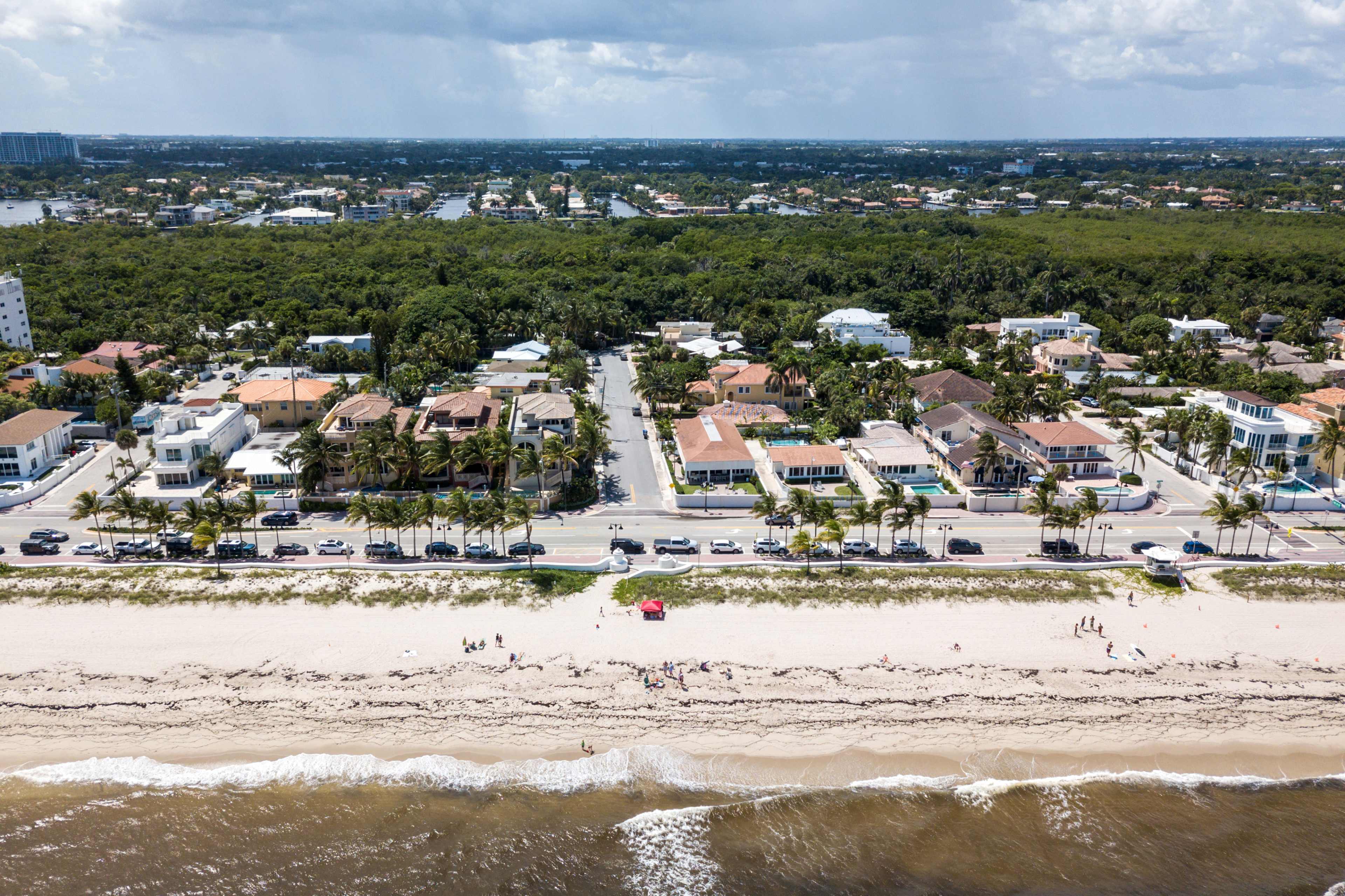An aerial view captures a beachside neighborhood with palm trees lining the road, residential houses on one side, and a sandy beach on the other.