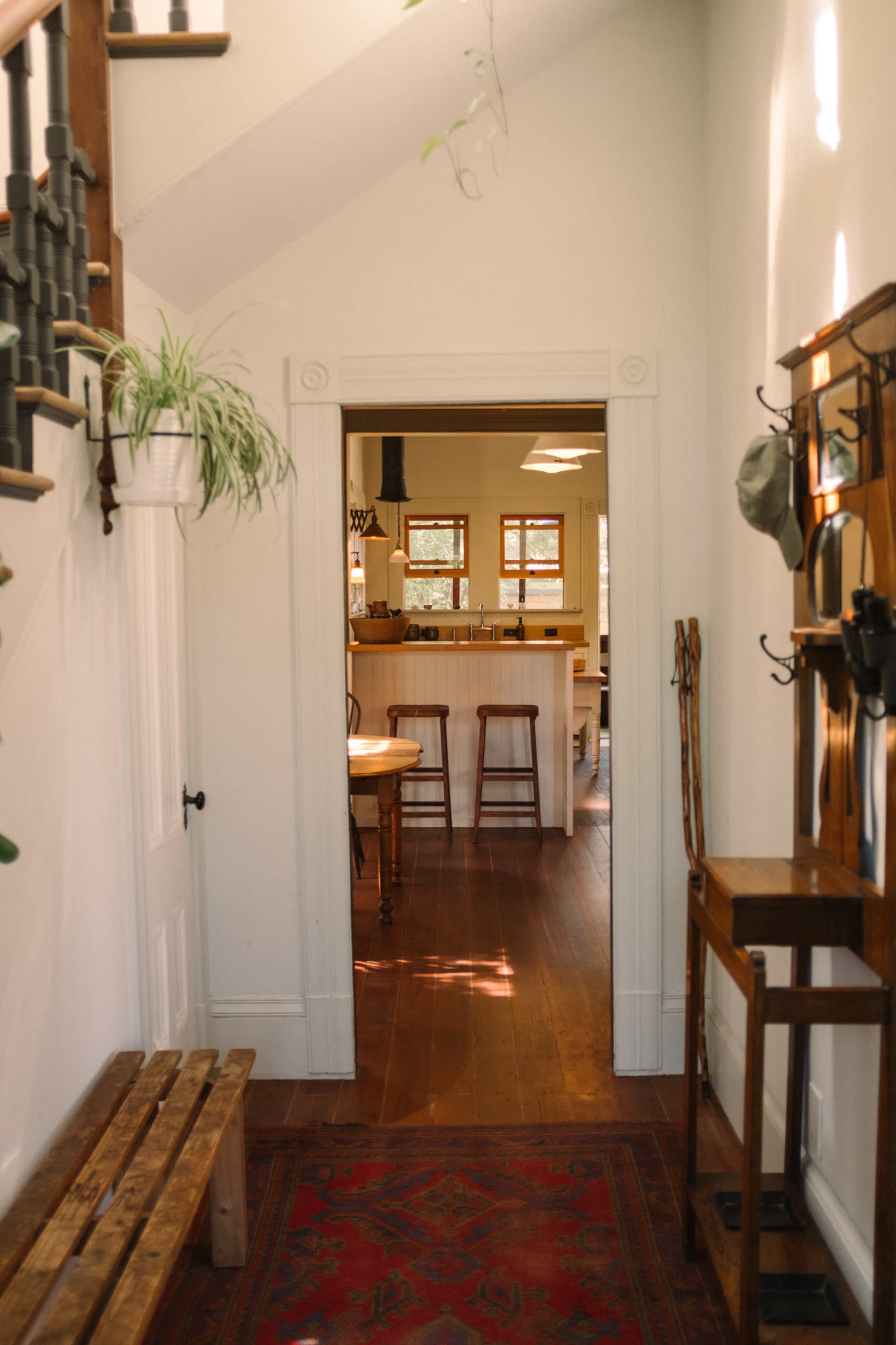 The image shows a hallway leading to a kitchen, featuring a wooden bench, coat rack, and light streaming through the windows.