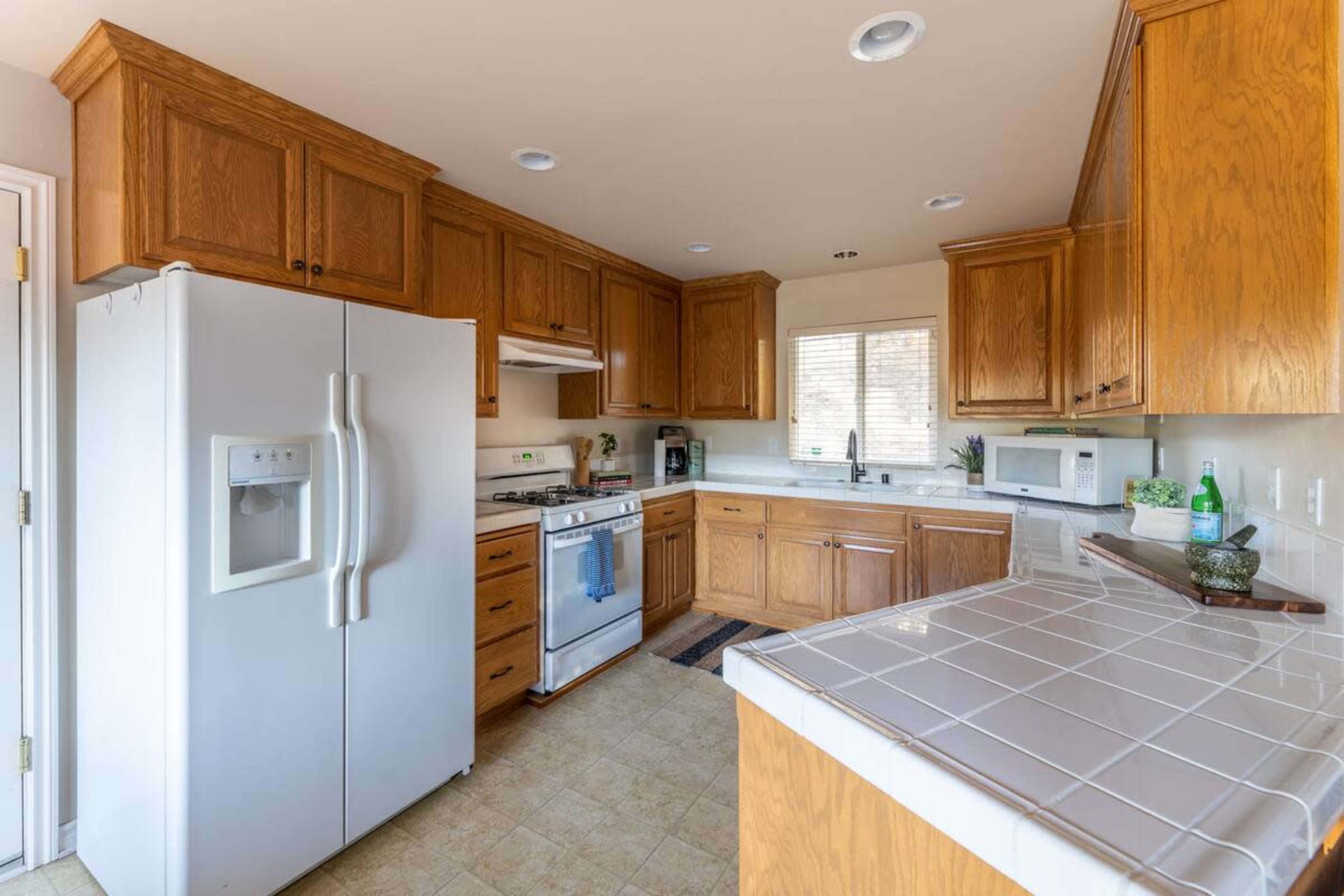 The image shows a kitchen with wooden cabinets, a white refrigerator, a stove, and a countertop with tile and appliances.