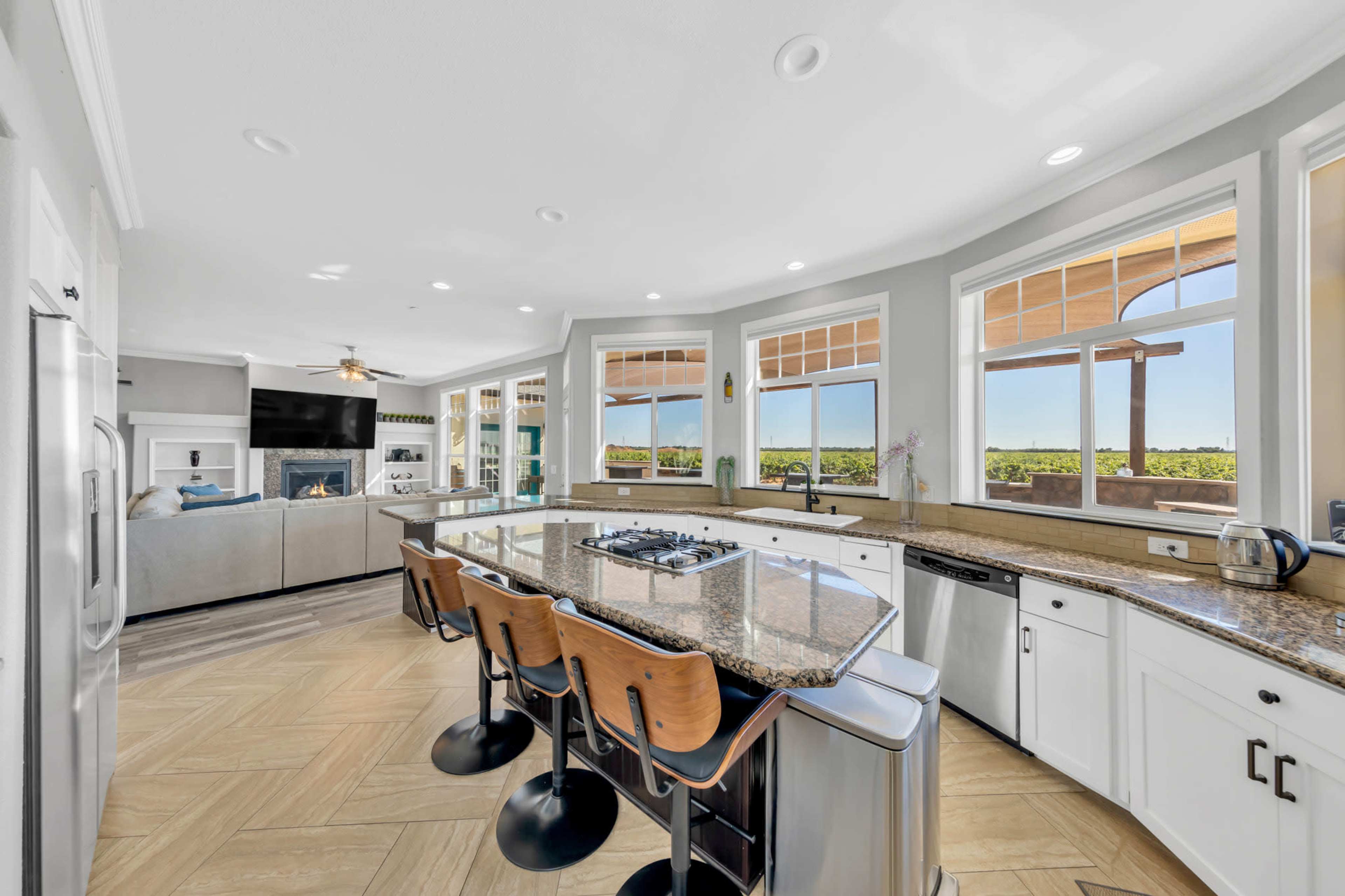 A modern kitchen with a large island, equipped with bar stools, overlooking a spacious living area and scenic outdoor views through multiple windows.