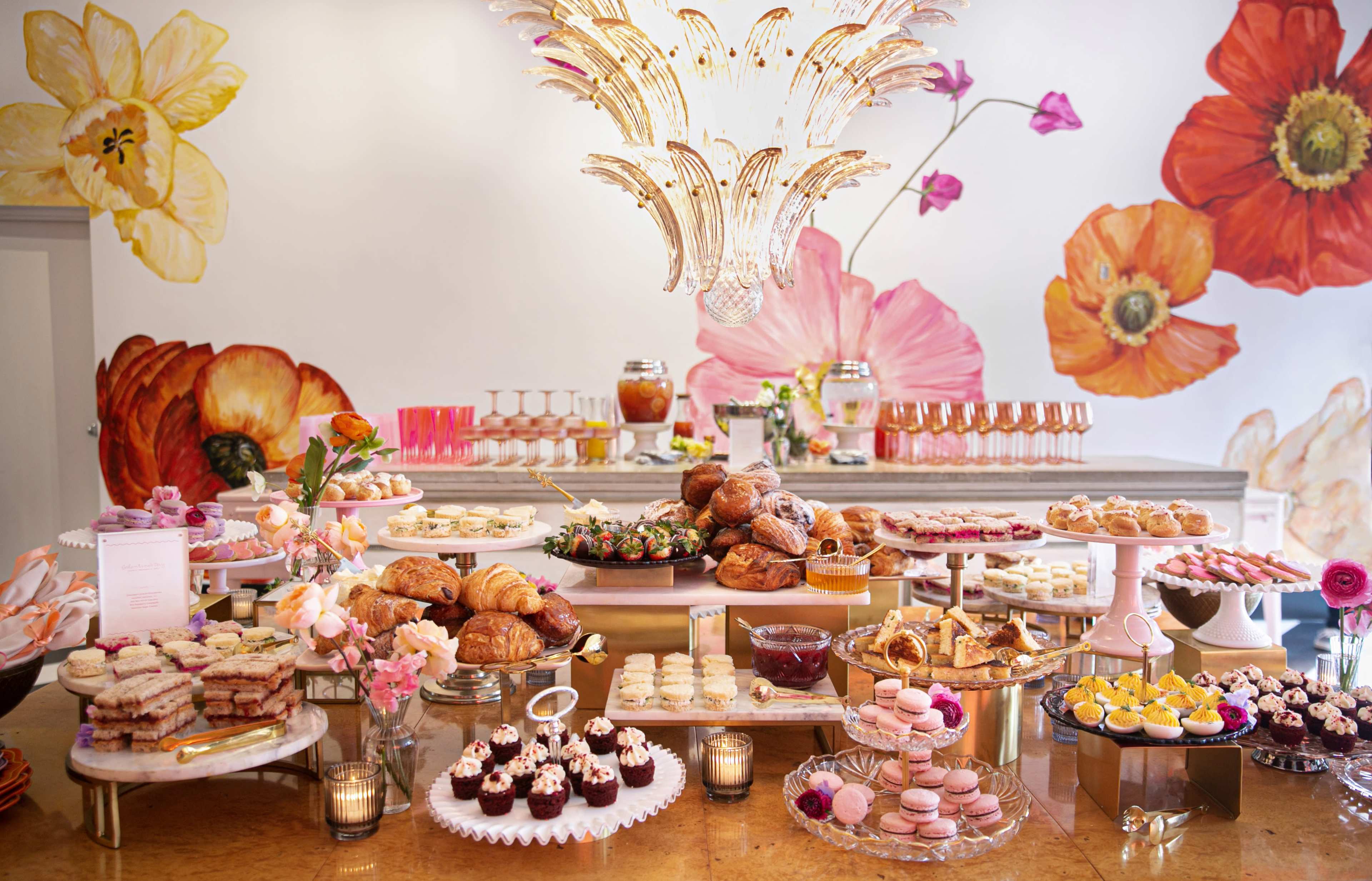 A lavish dessert table is arranged with an assortment of pastries, cakes, and decorative drinks beneath a floral mural.