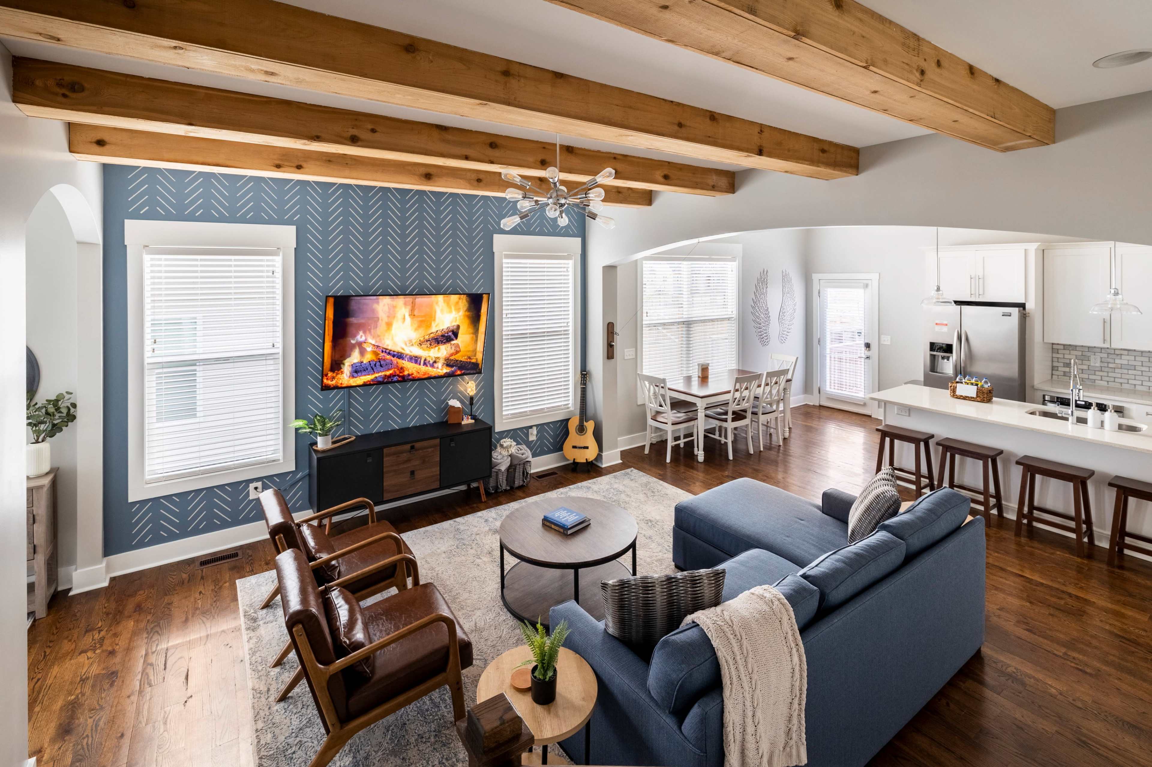 A spacious living room features a blue sectional couch, wooden beams on the ceiling, a television mounted above a dark cabinet, and a cozy fireplace.