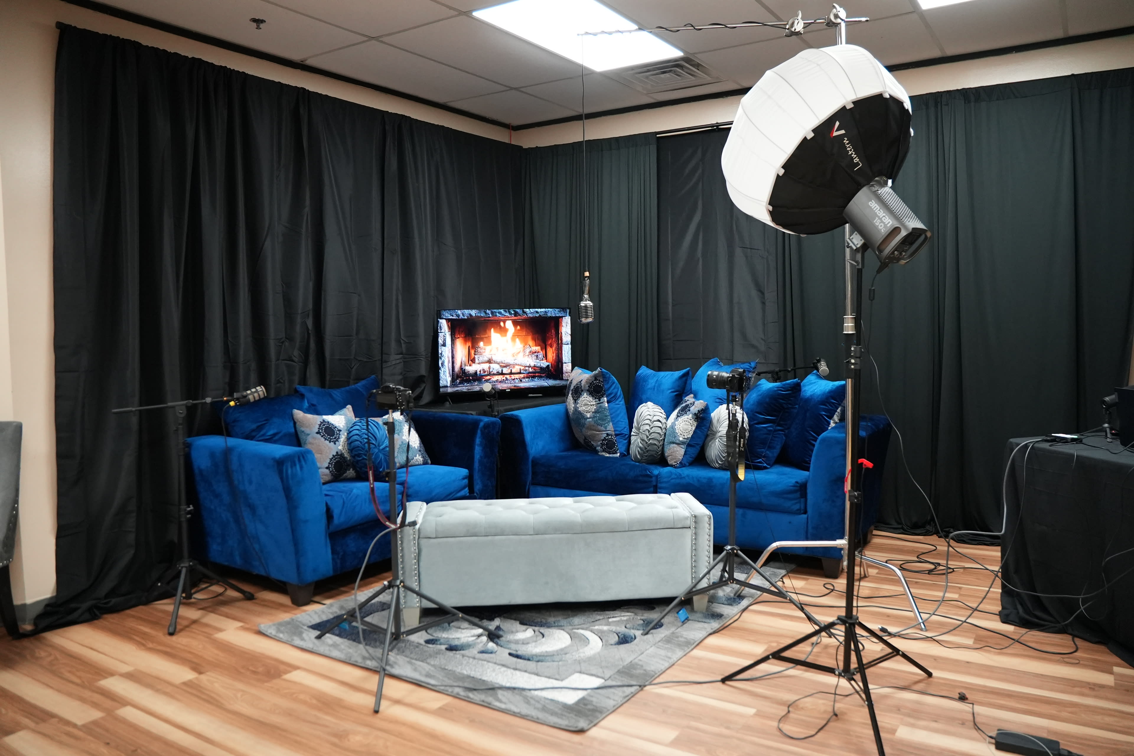 The image shows a styled indoor setting with blue velvet sofas arranged around a decorative coffee table, a television displaying a fireplace, and lighting equipment set up in the background.
