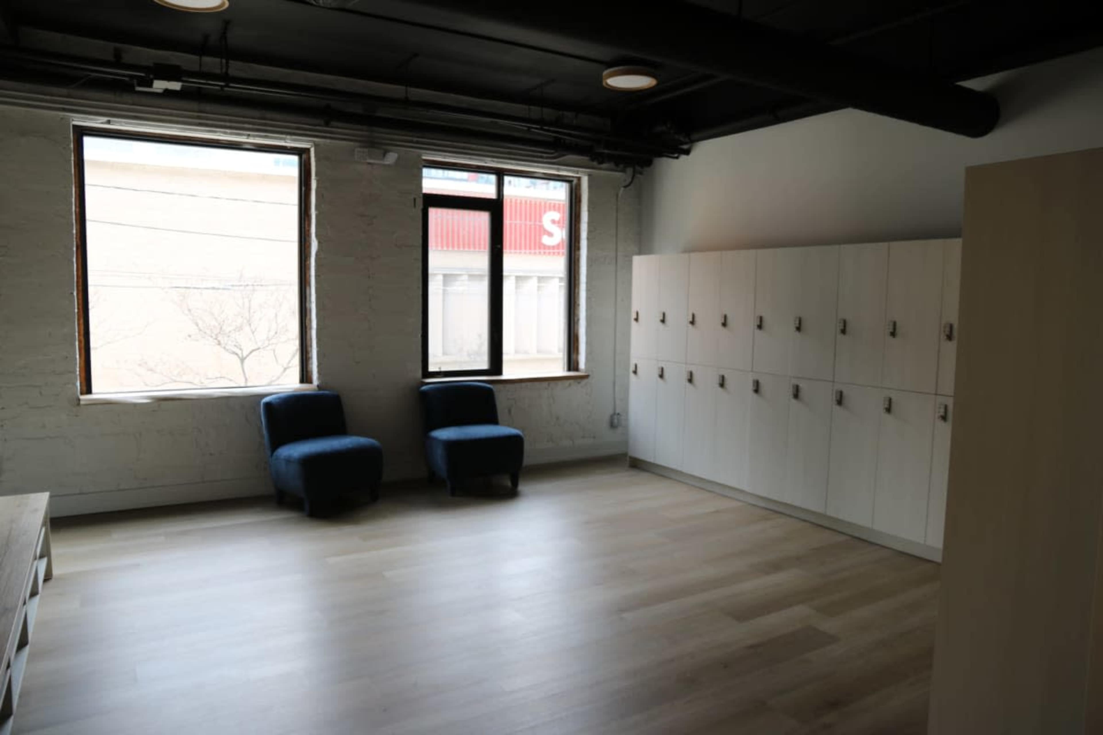 The image shows a minimalistic room with two blue chairs, a row of white lockers, and large windows letting in natural light.