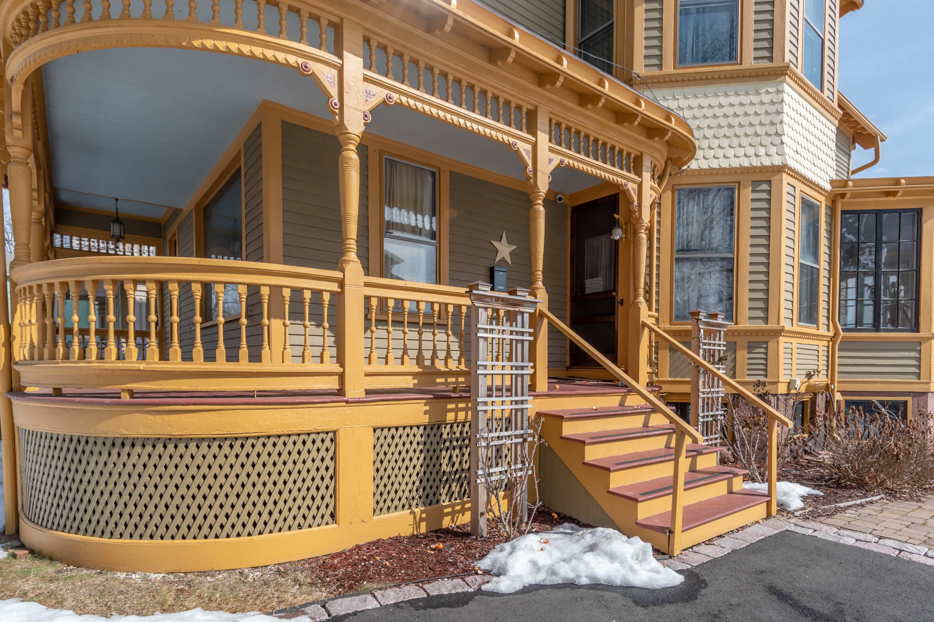 A Victorian-style house with a large, intricately designed porch, a set of stairs leading to the entrance, and patches of snow on the ground.