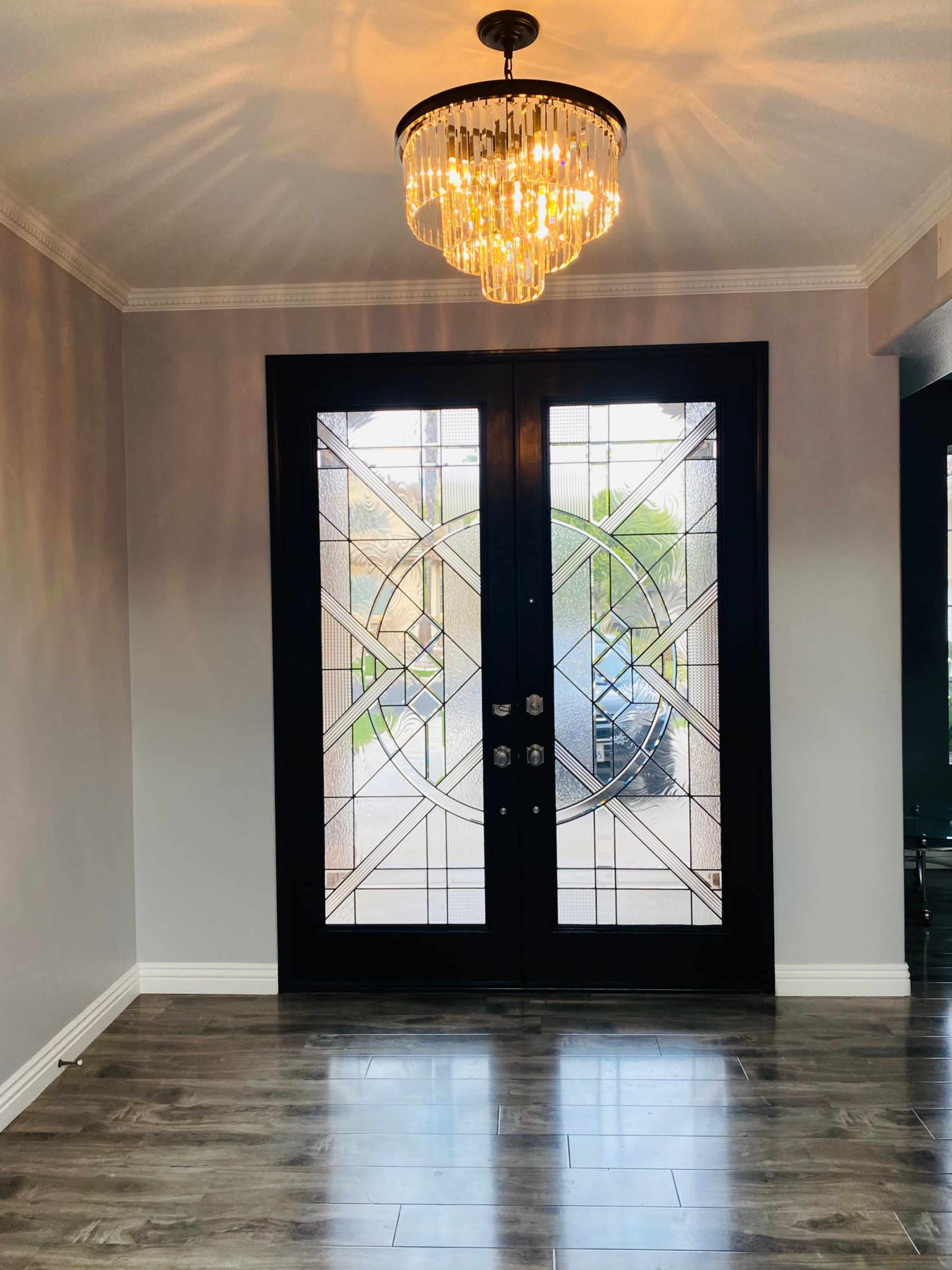 An entryway with double glass doors featuring geometric designs, complemented by a chandelier above and wooden flooring beneath.