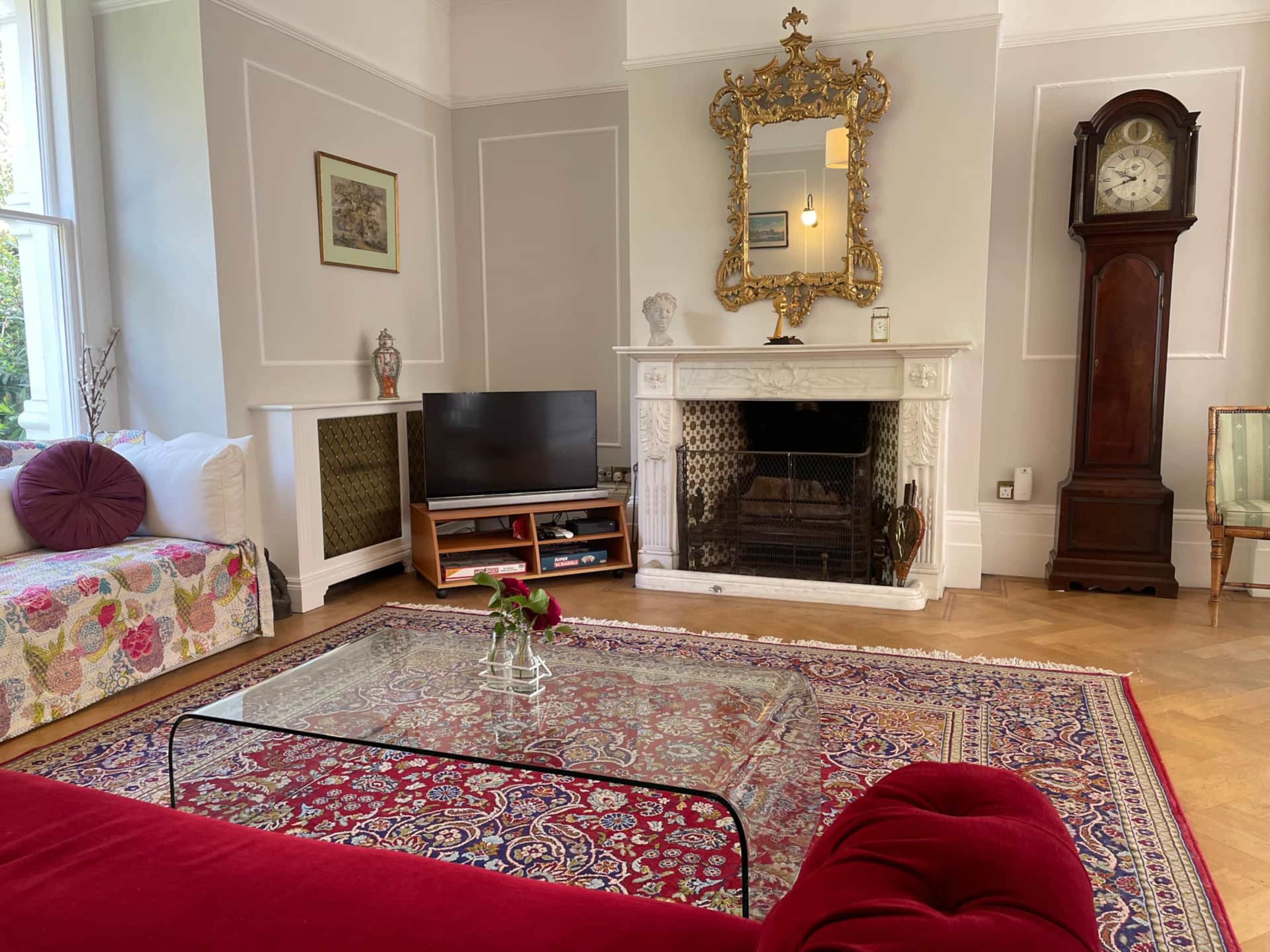 The room features a patterned sofa, a television on a stand, a large ornate mirror above a marble fireplace, a vintage clock, and a glass coffee table on a decorative rug.