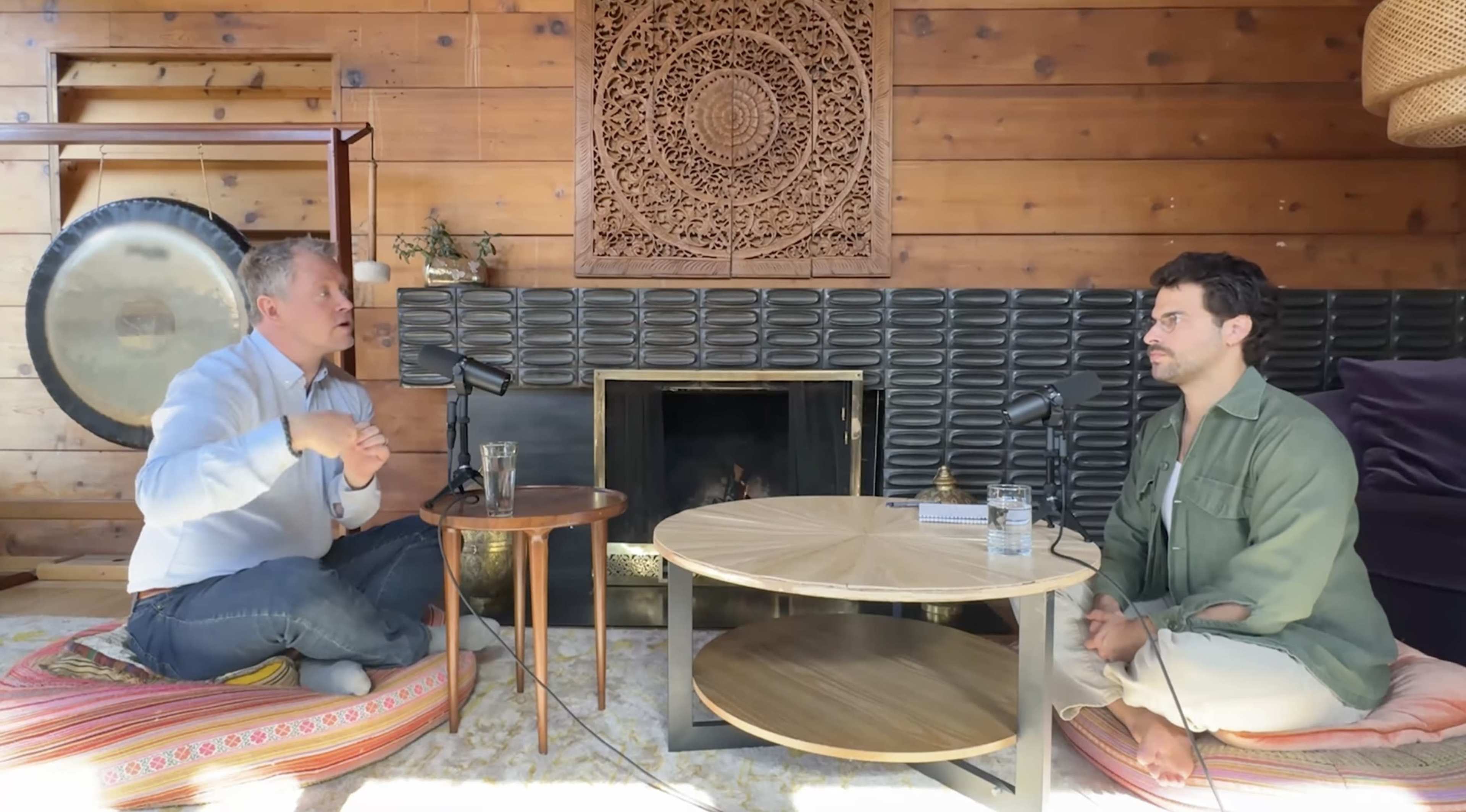 Two men sit across from each other in a wooden room, discussing with microphones placed on small tables between them, and a decorative wall panel in the background.