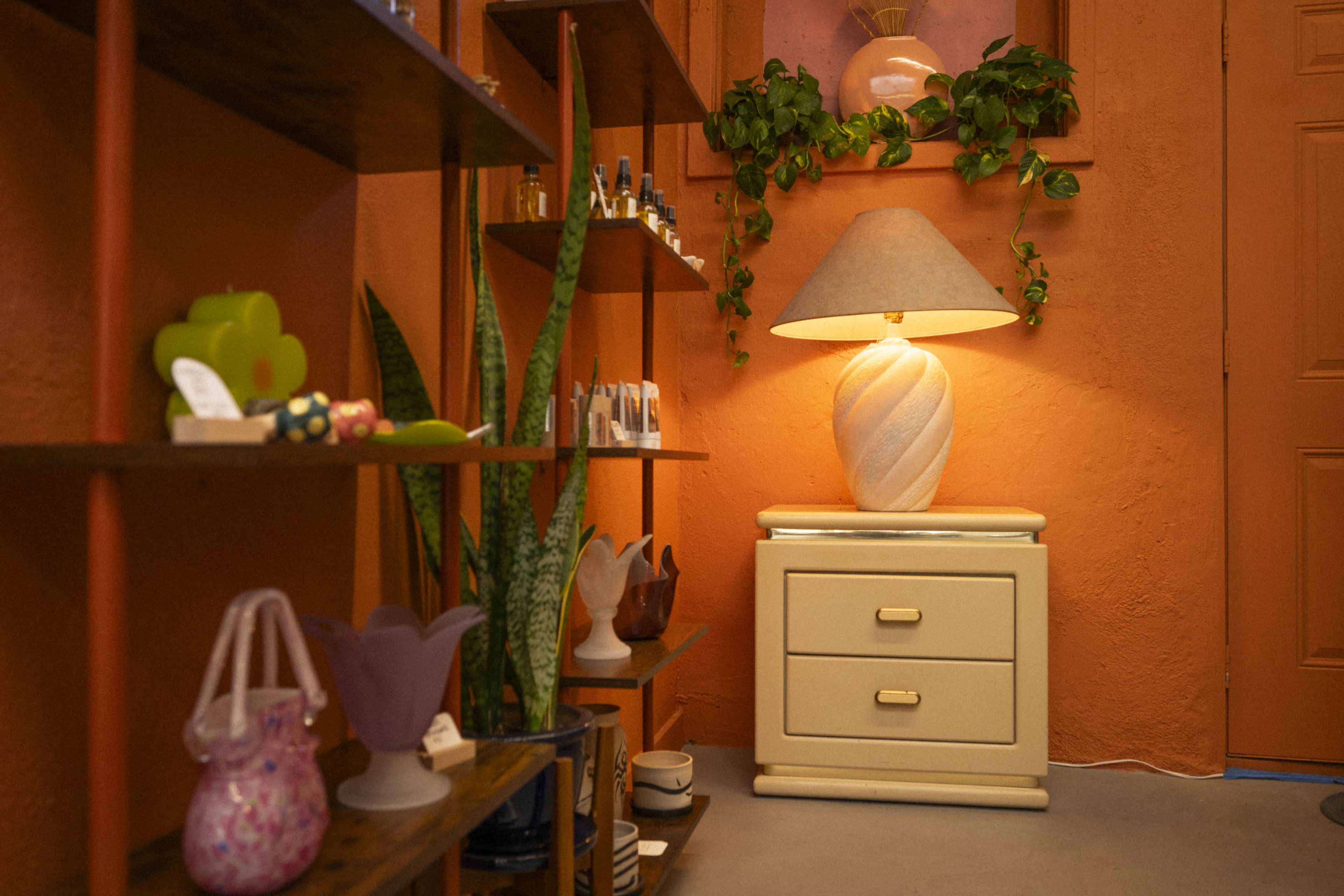 The scene features a cozy interior showcasing a shelf filled with various colorful decorative items beside a nightstand with a lamp and a potted plant against an orange wall.
