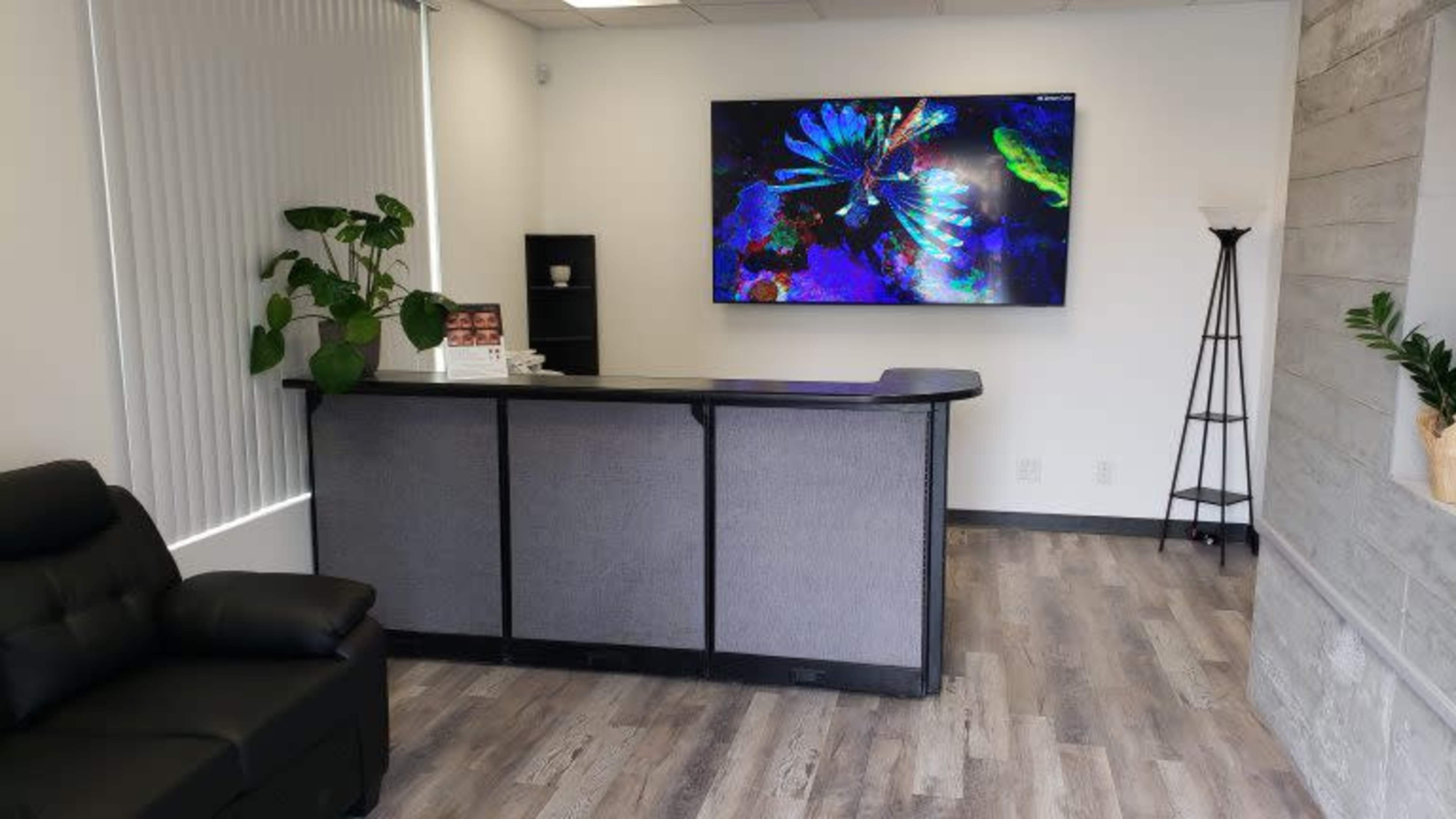 The image shows a modern reception area featuring a gray front desk, a large wall-mounted screen displaying colorful artwork, and a comfortable black couch.