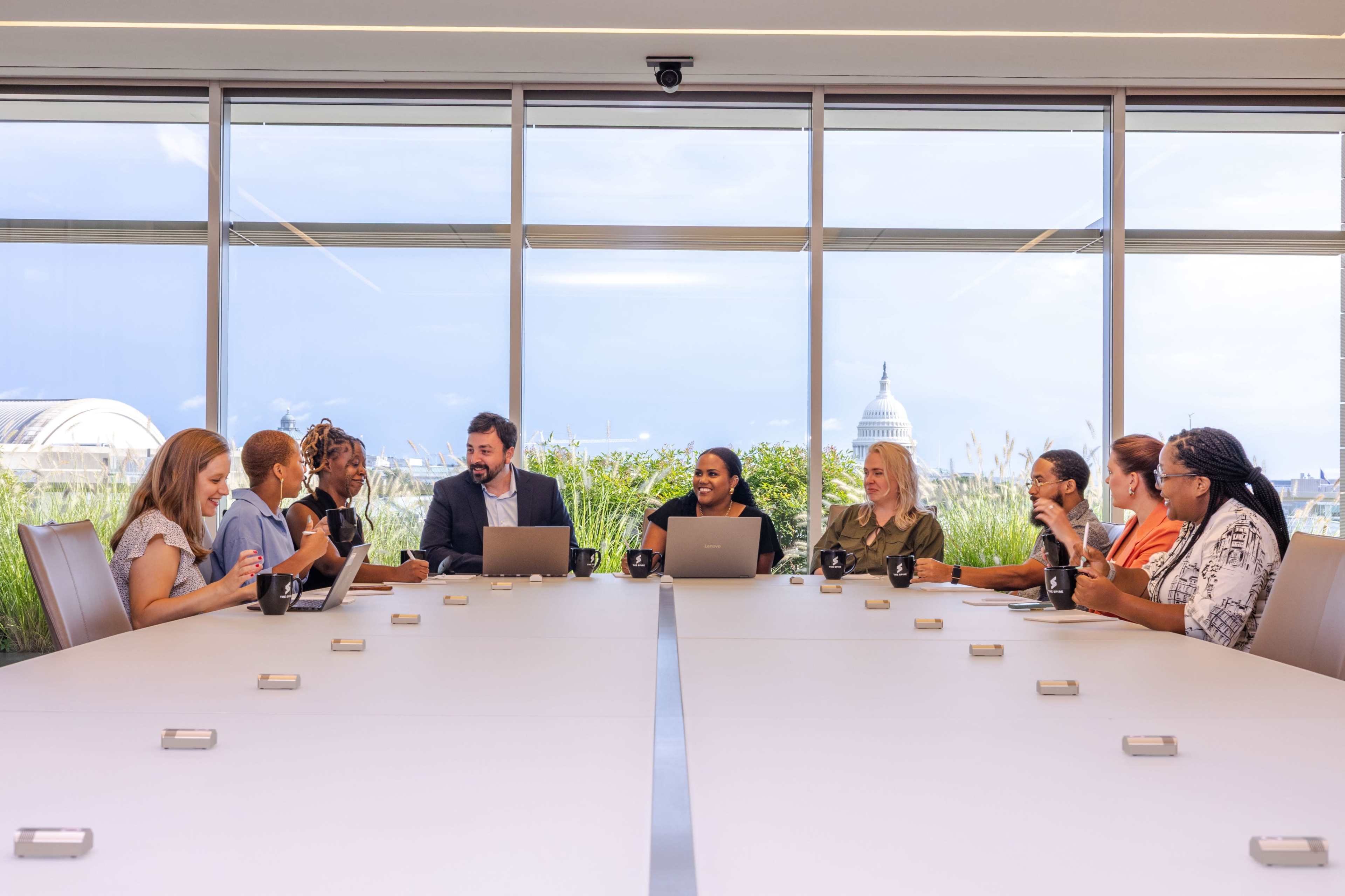A group of diverse professionals is seated around a large conference table, engaged in discussion, with a view of a city skyline visible through the windows.