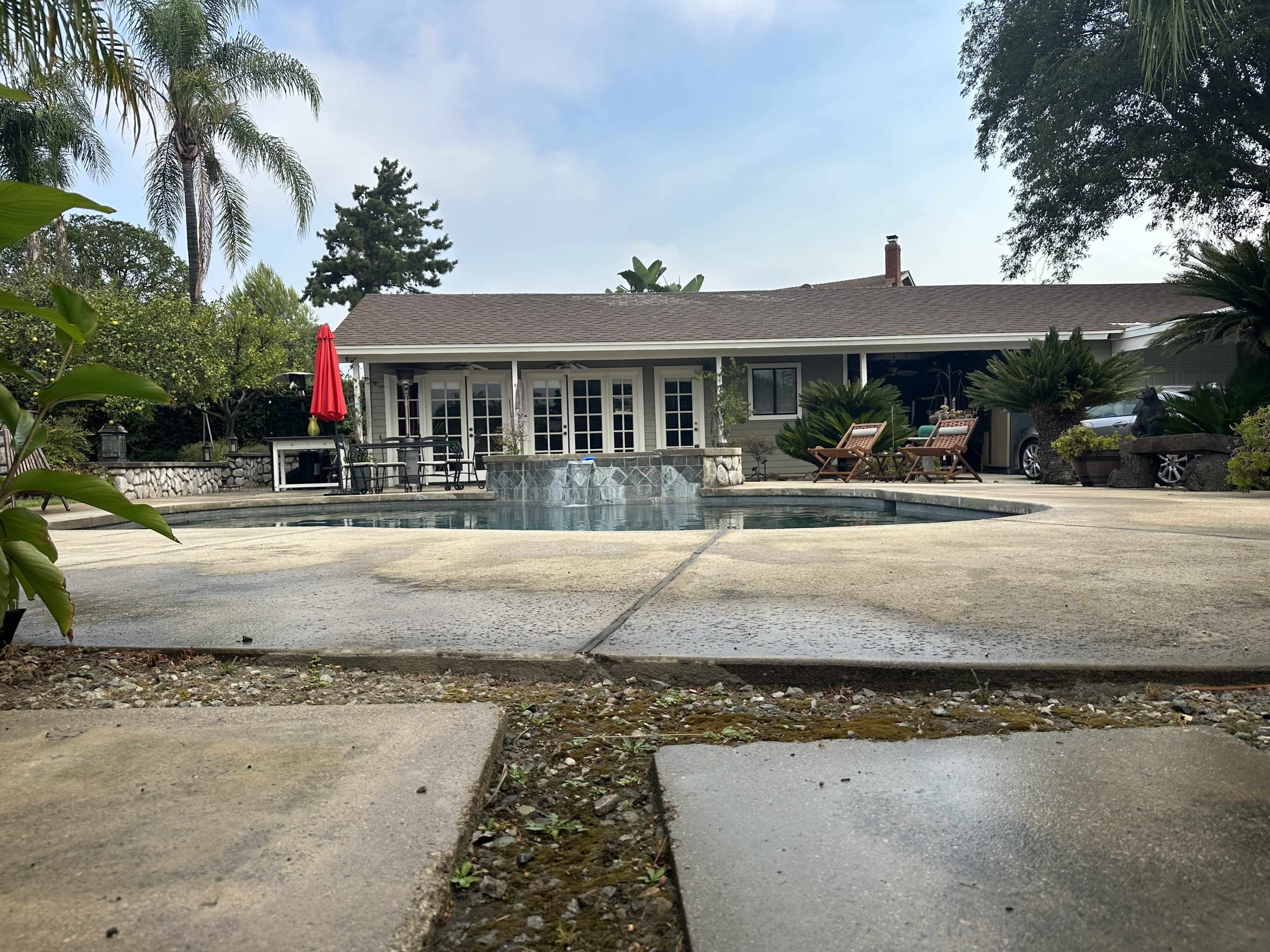 A clear swimming pool surrounded by stone flooring and tropical plants, with a house featuring large windows and a red umbrella in the background.