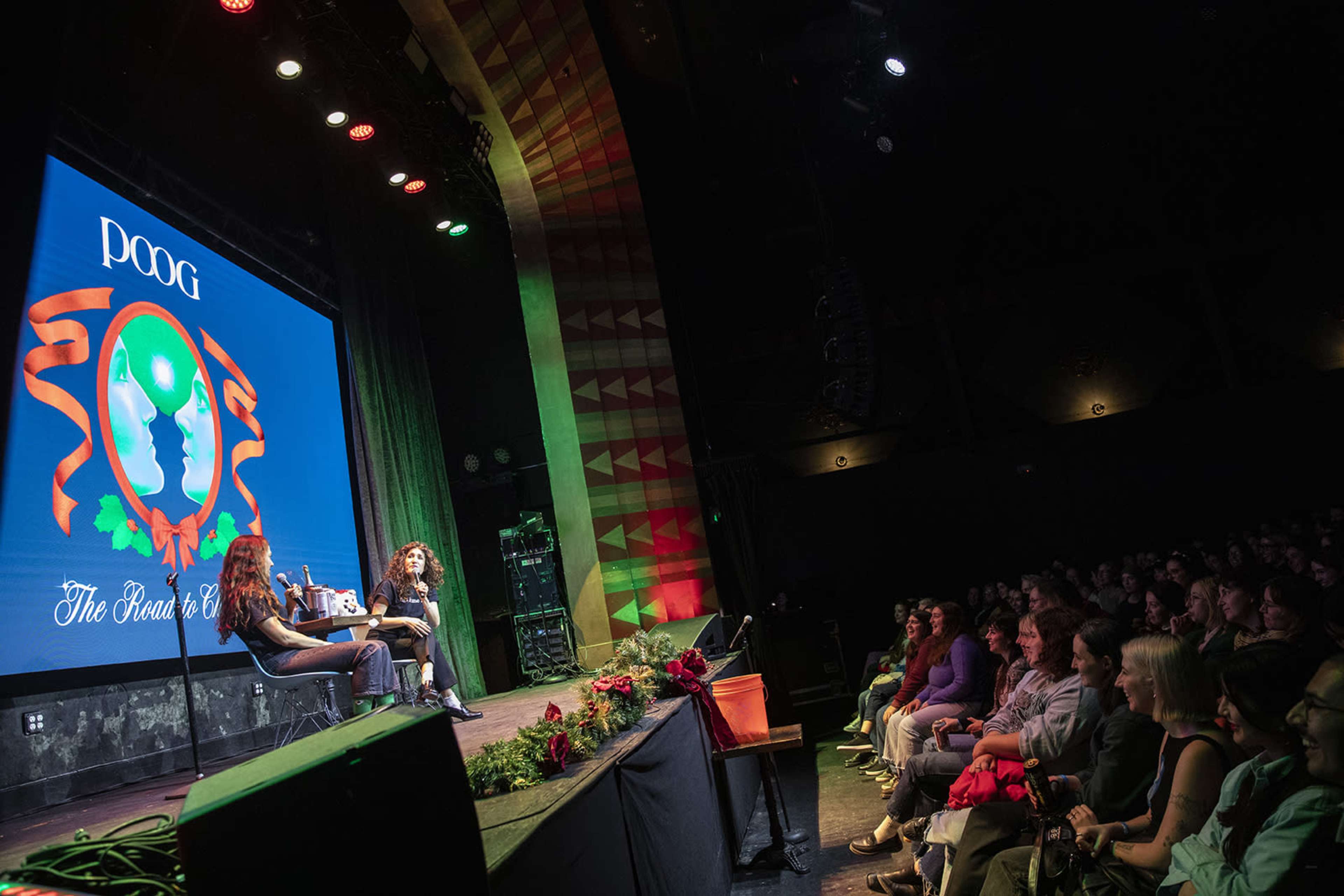 A stage with two speakers discussing a topic, while an audience listens attentively in a theater setting.