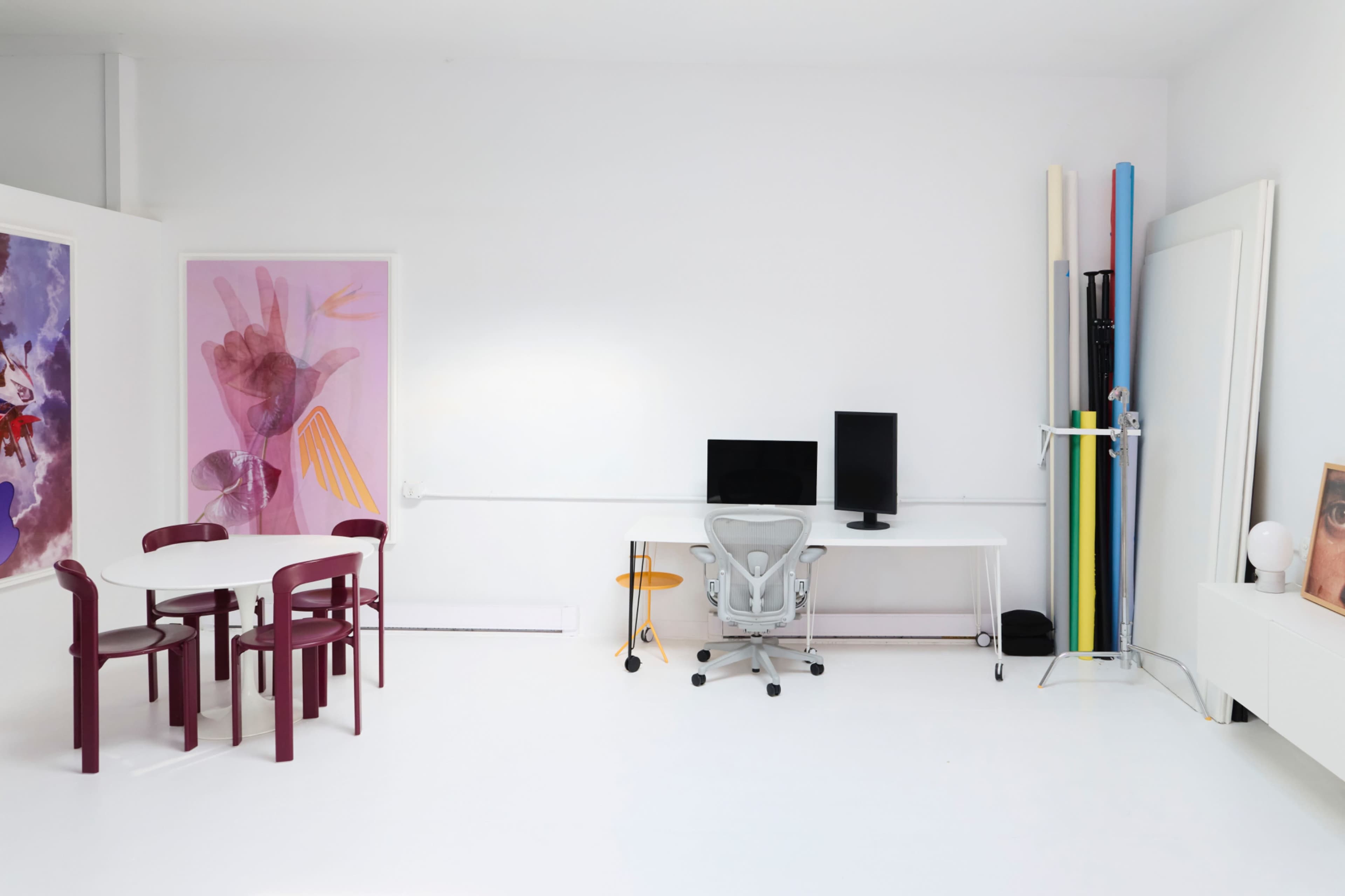 The image shows a bright, minimalist workspace featuring a white interior with a desk, two monitors, a white chair, a small table with four maroon chairs, and art materials in the corner.