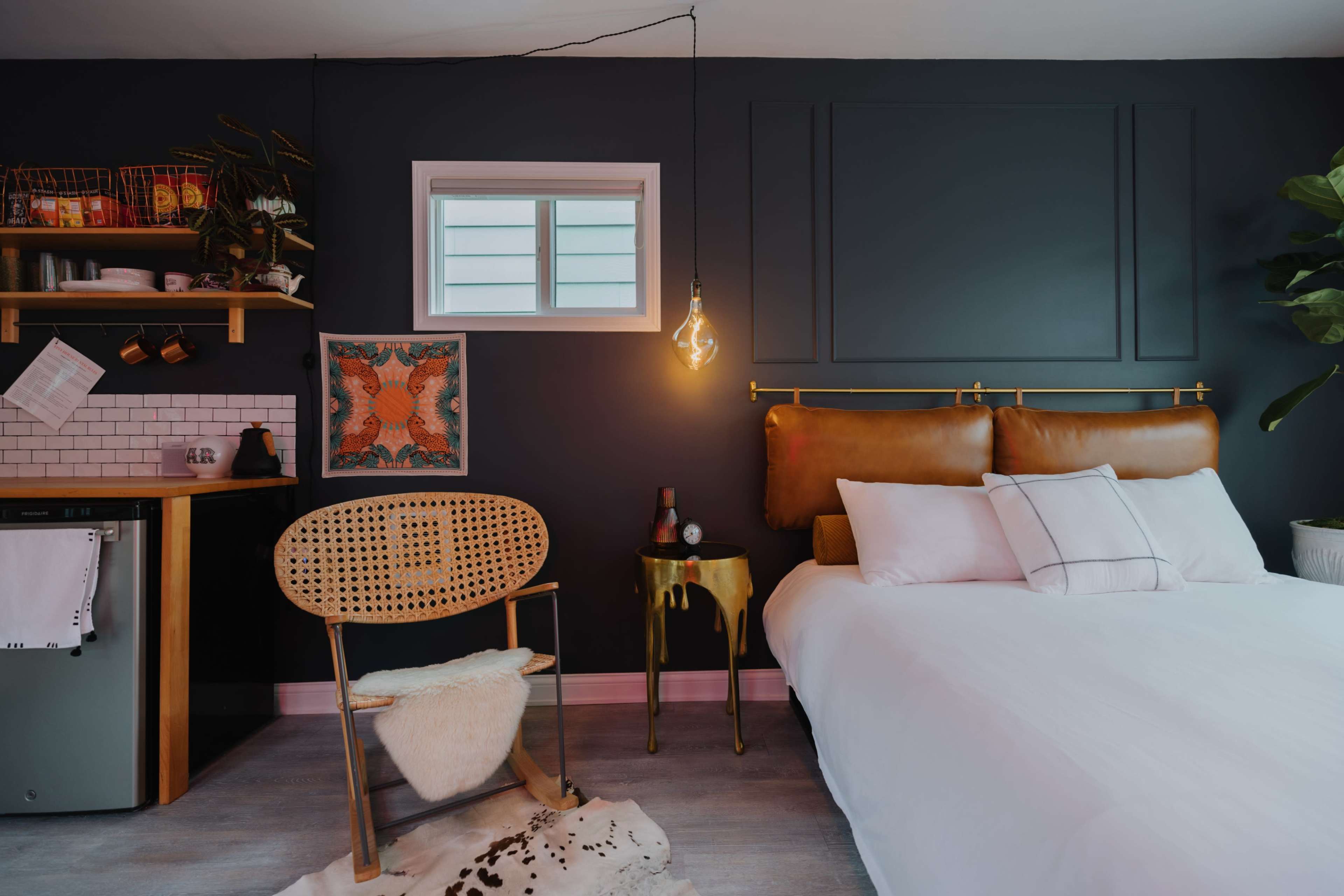 The image shows a modern bedroom featuring a bed with white linens, a rattan chair, a small kitchen area with a mini fridge, and a wall with dark paint and a decorative piece.