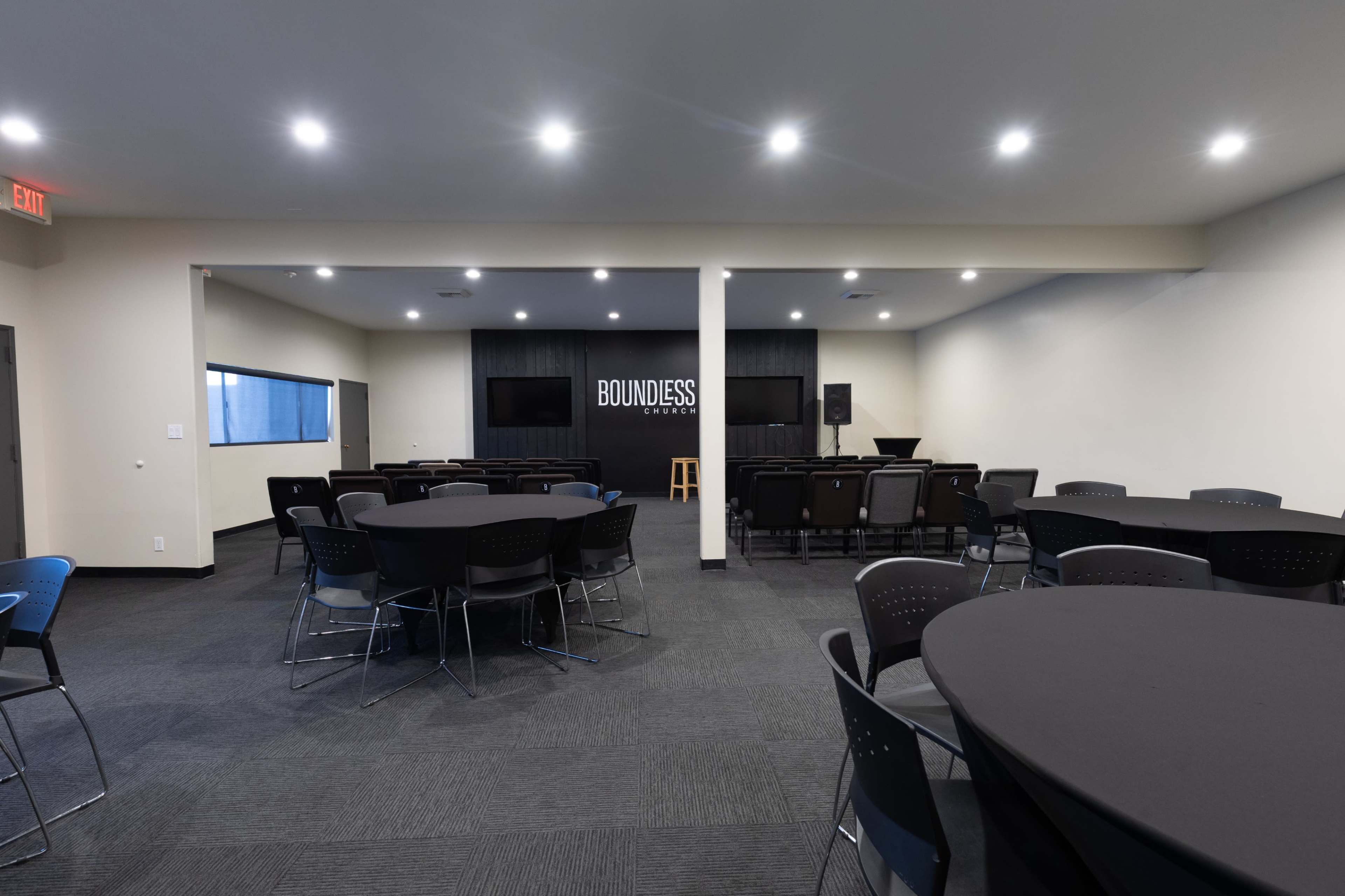 The image shows a spacious, well-lit event room featuring round tables with black tablecloths and a stage with the word "BOUNDLESS" displayed on a dark wall.