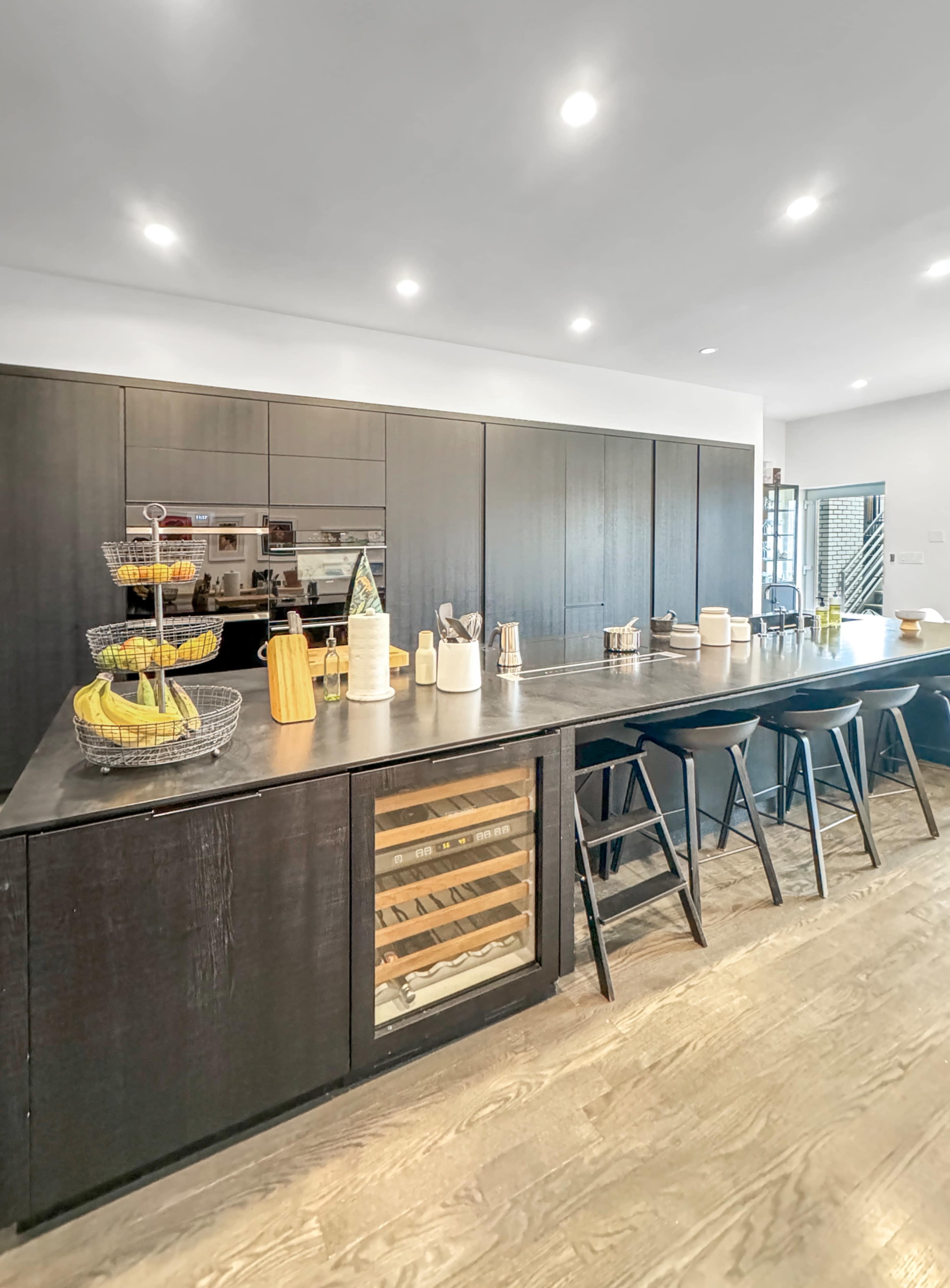 The image shows a modern kitchen featuring dark cabinetry, a long island with bar stools, and organized storage on the countertop.