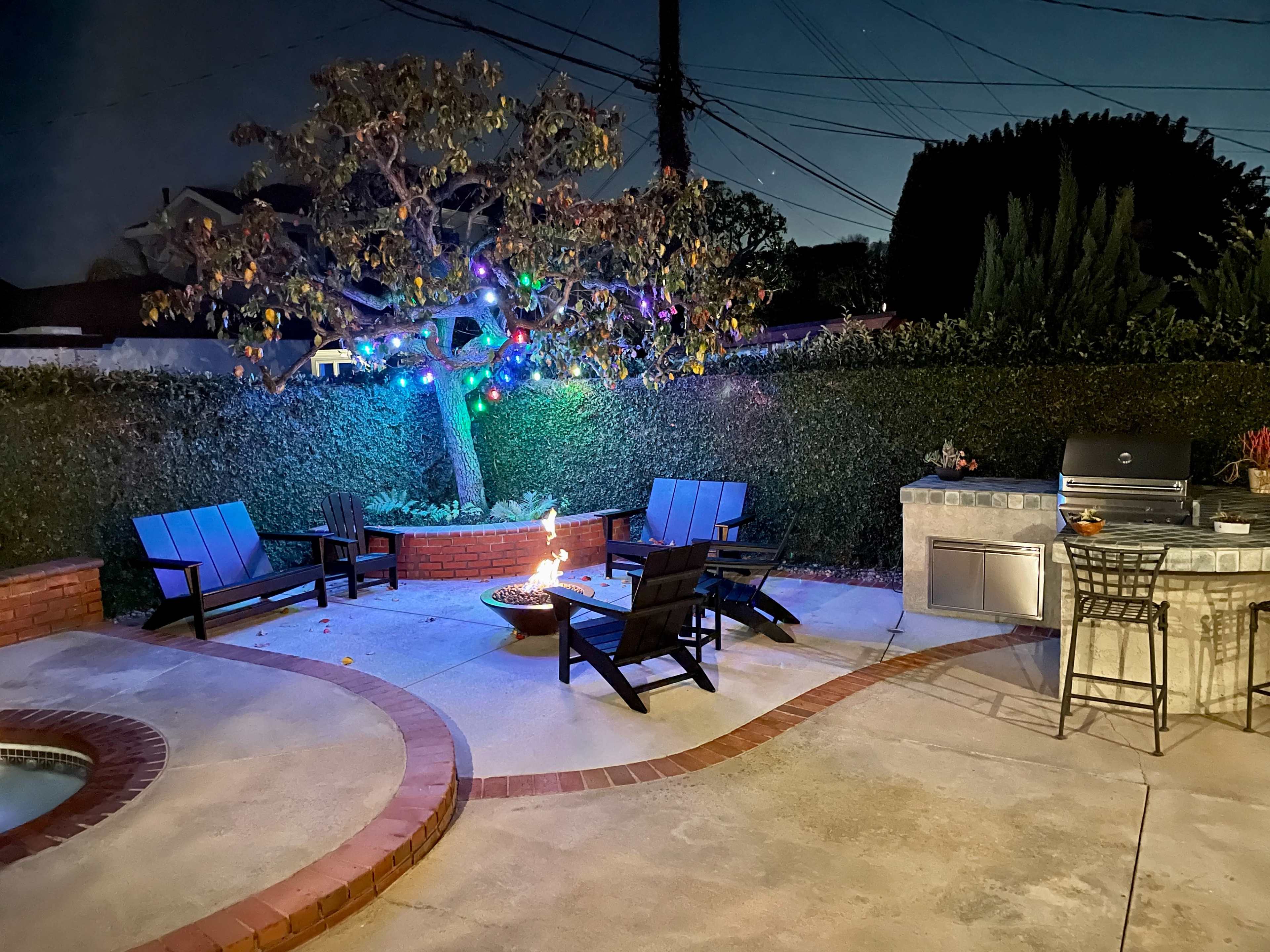 A backyard patio features several black chairs arranged around a fire pit, a BBQ grill, and a tree adorned with colorful lights amid a landscaped area.