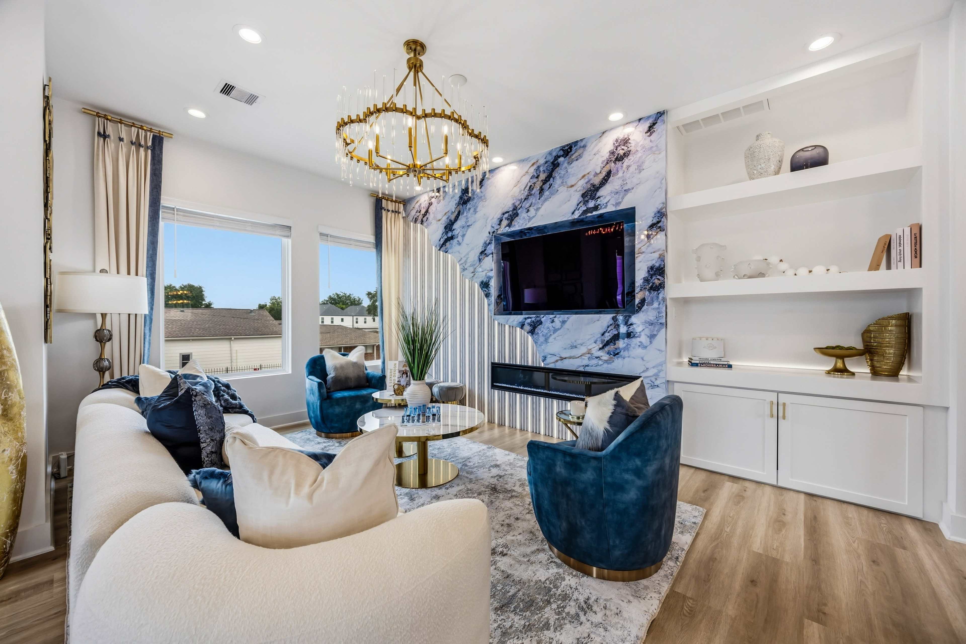 The living room features a modern design with a curved white sofa, two blue accent chairs, a round coffee table, and a wall adorned with blue and white marble, complemented by a chandelier and a built-in shelving unit.