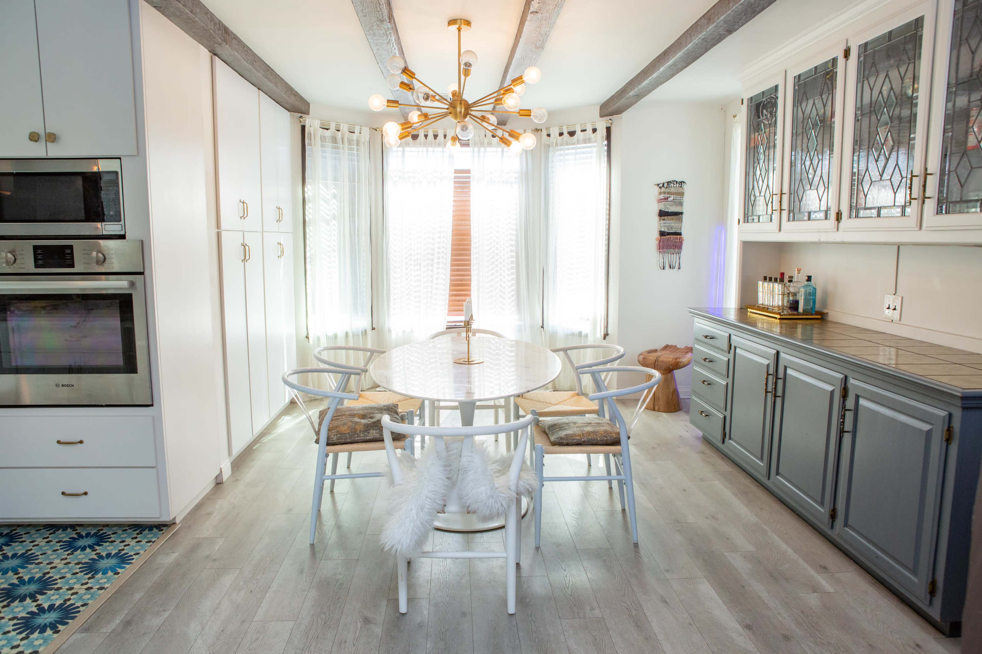 A spacious dining area features a round table surrounded by white chairs, illuminated by a modern chandelier, with large windows allowing natural light to enter.