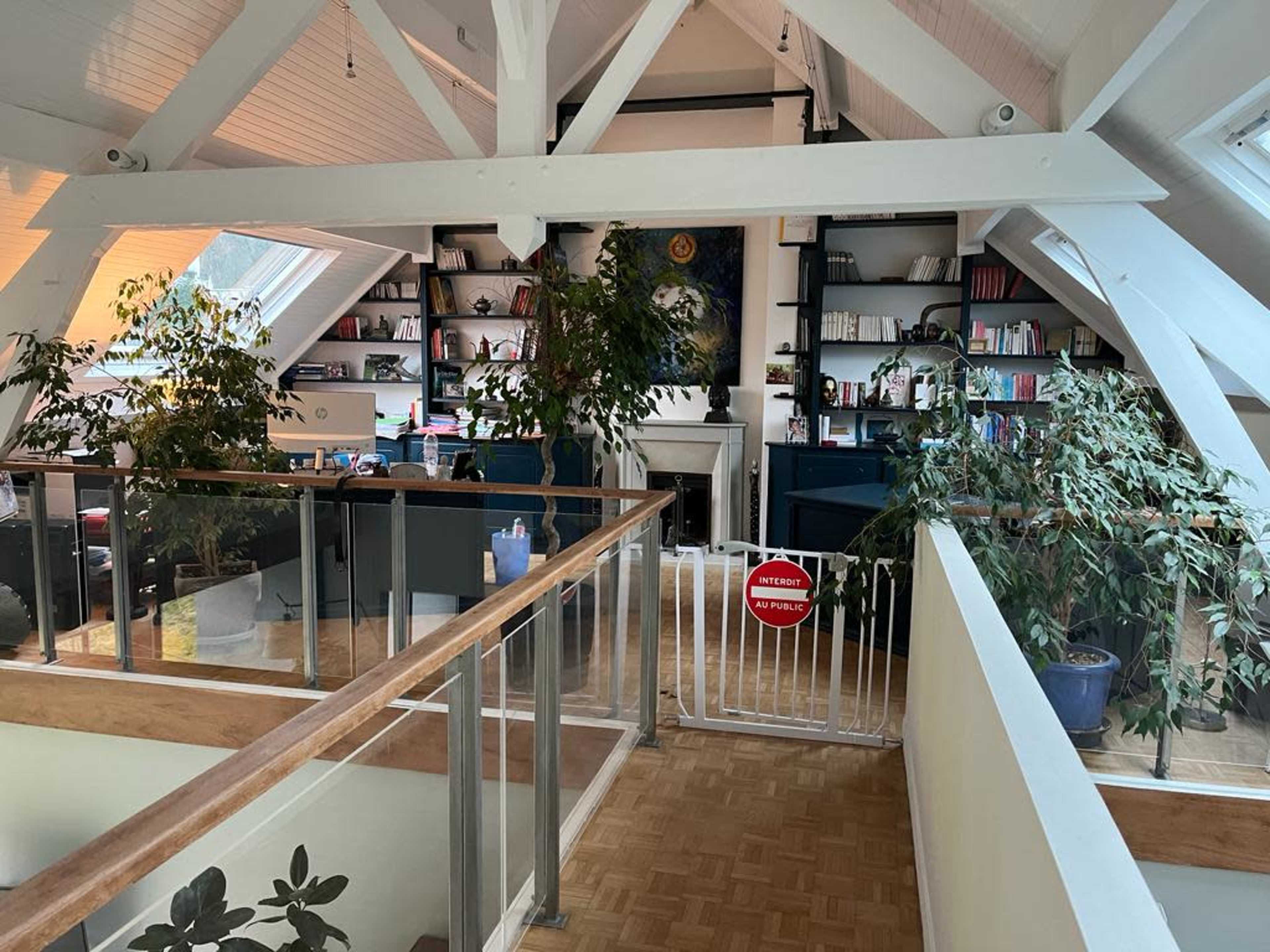 The image shows a spacious, bright room with a wooden railing, bookshelves filled with books, and several plants distributed throughout the space.