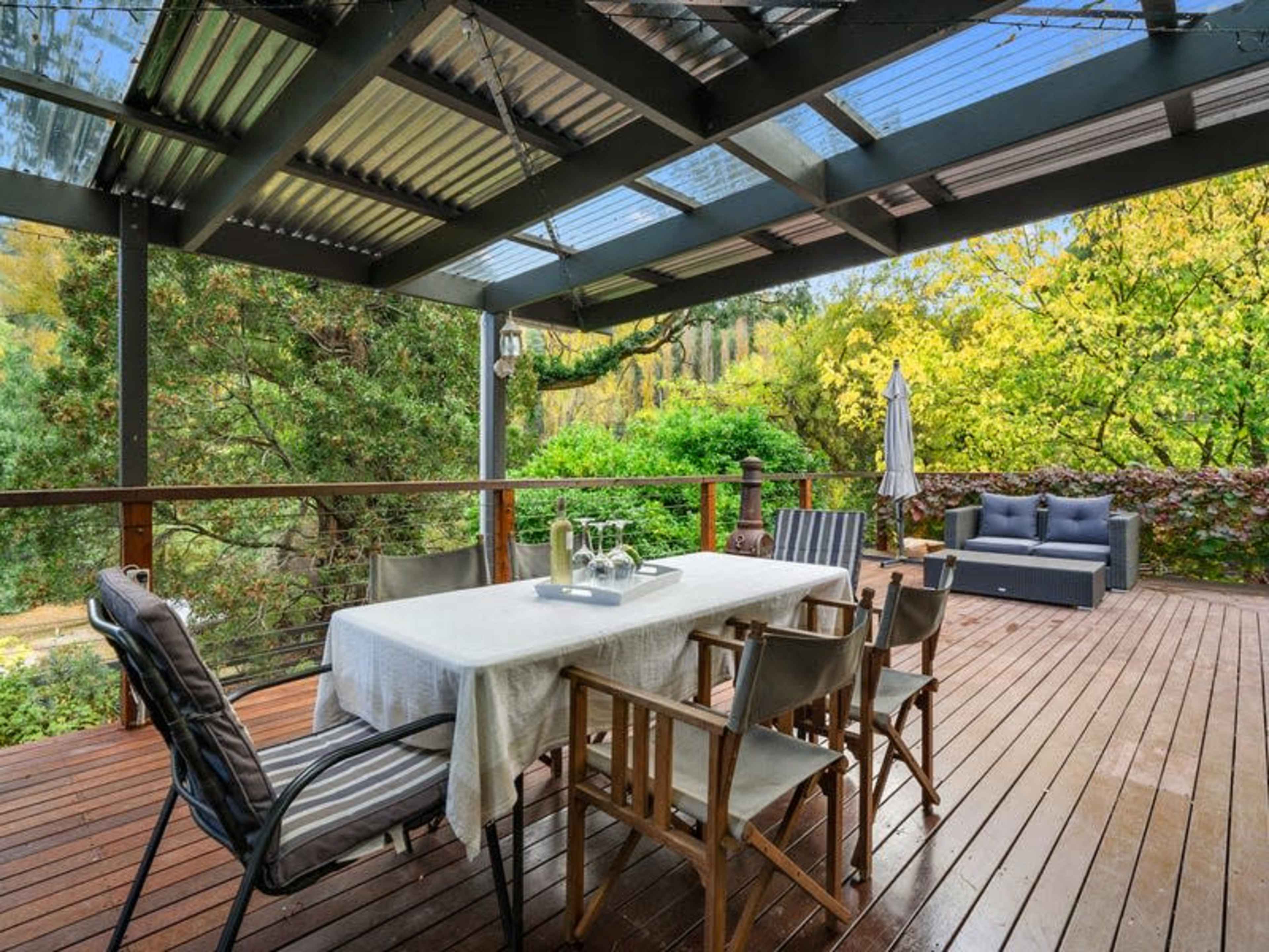 A wooden deck features a dining table surrounded by chairs, with a backdrop of lush greenery and a seating area.