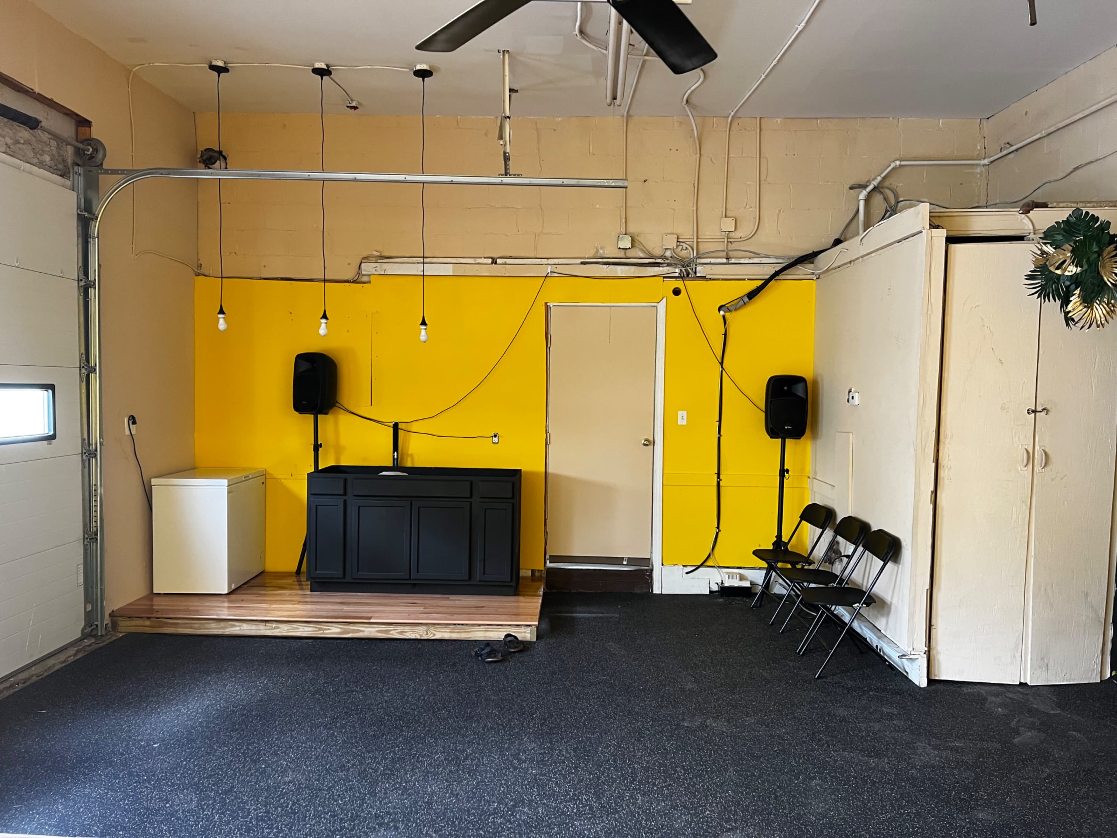 The image shows a garage with a yellow wall, a black bar-style counter, a washing machine, speakers, a door, and a few folding chairs.