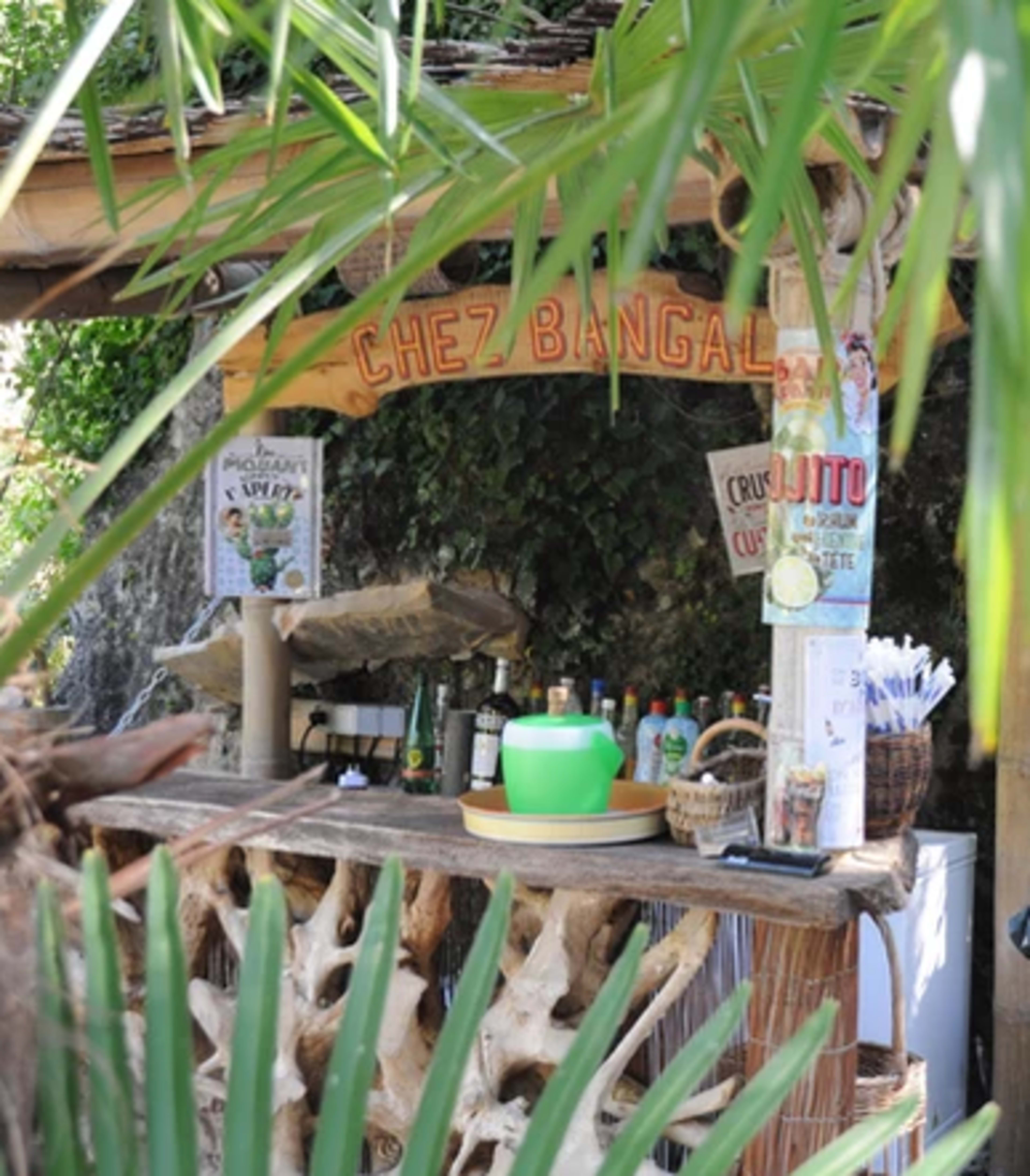 A rustic outdoor bar named "Chez Bangal" features a wooden counter and various bottles displayed behind it, surrounded by tropical greenery.