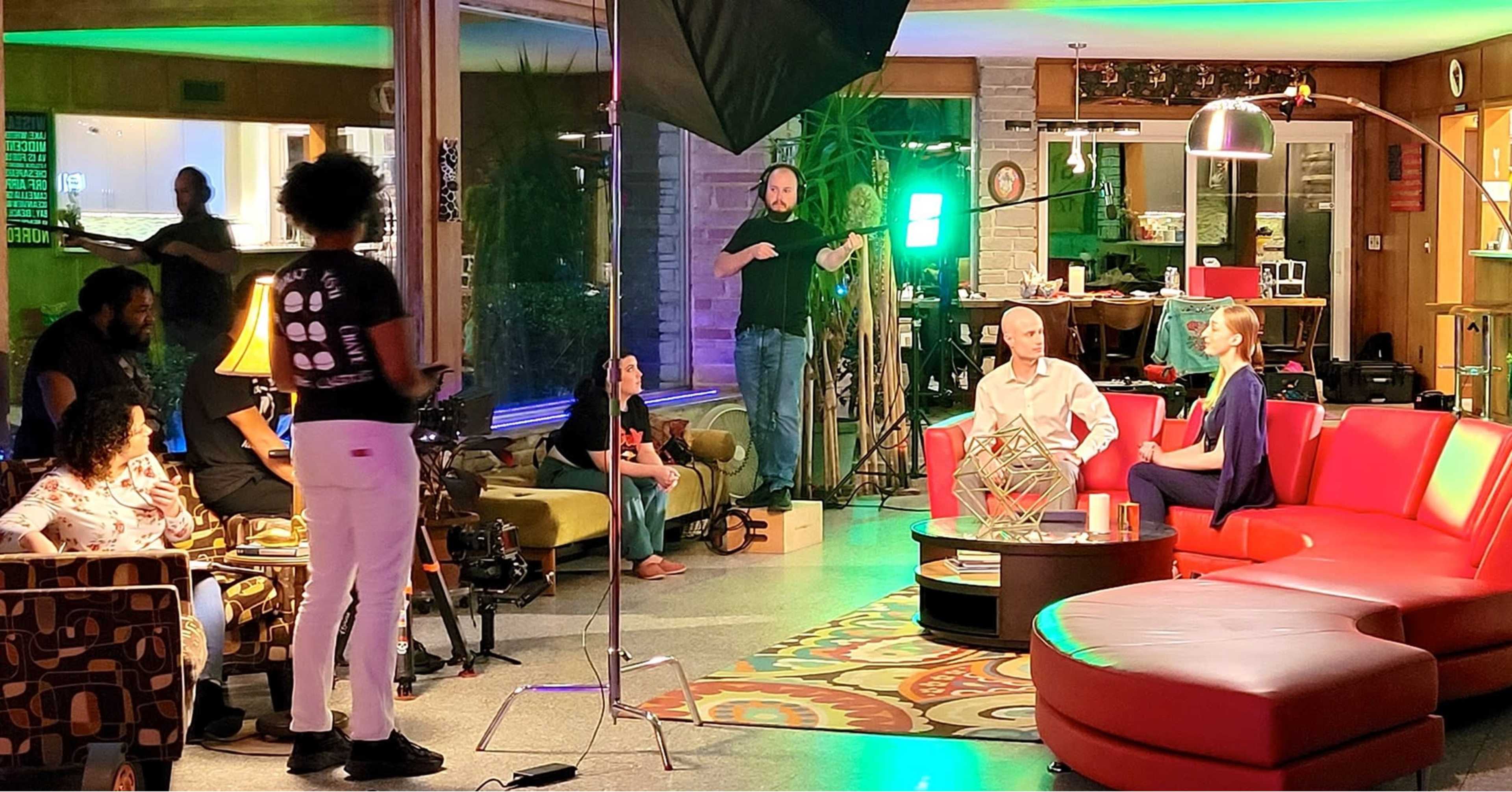 A film crew is setting up equipment for an interview scene in a modern indoor space featuring colorful furniture and decorative lighting.