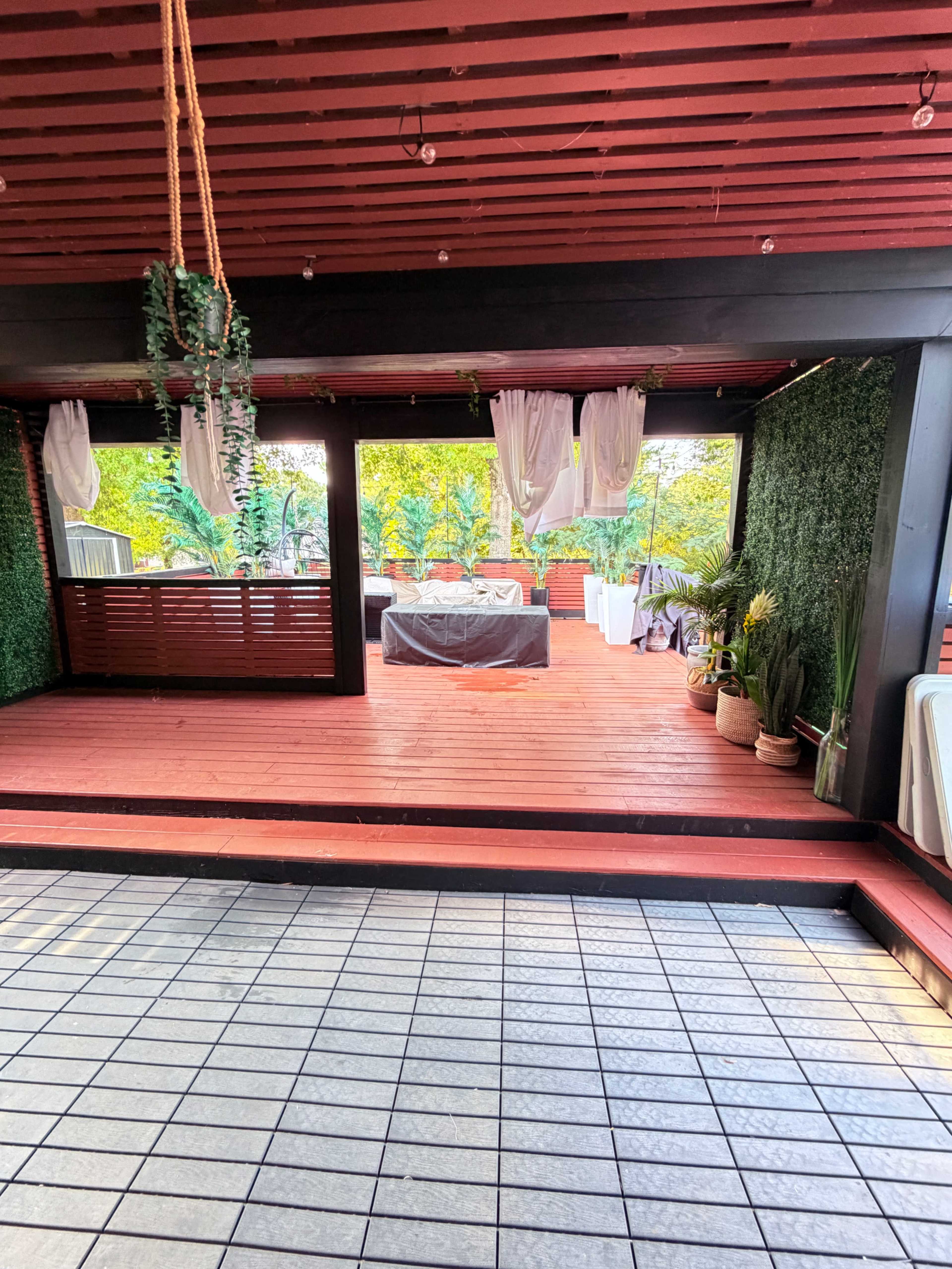 The image shows a spacious outdoor venue with wooden flooring, decorated with greenery and white drapes, and several tables set up for an event.