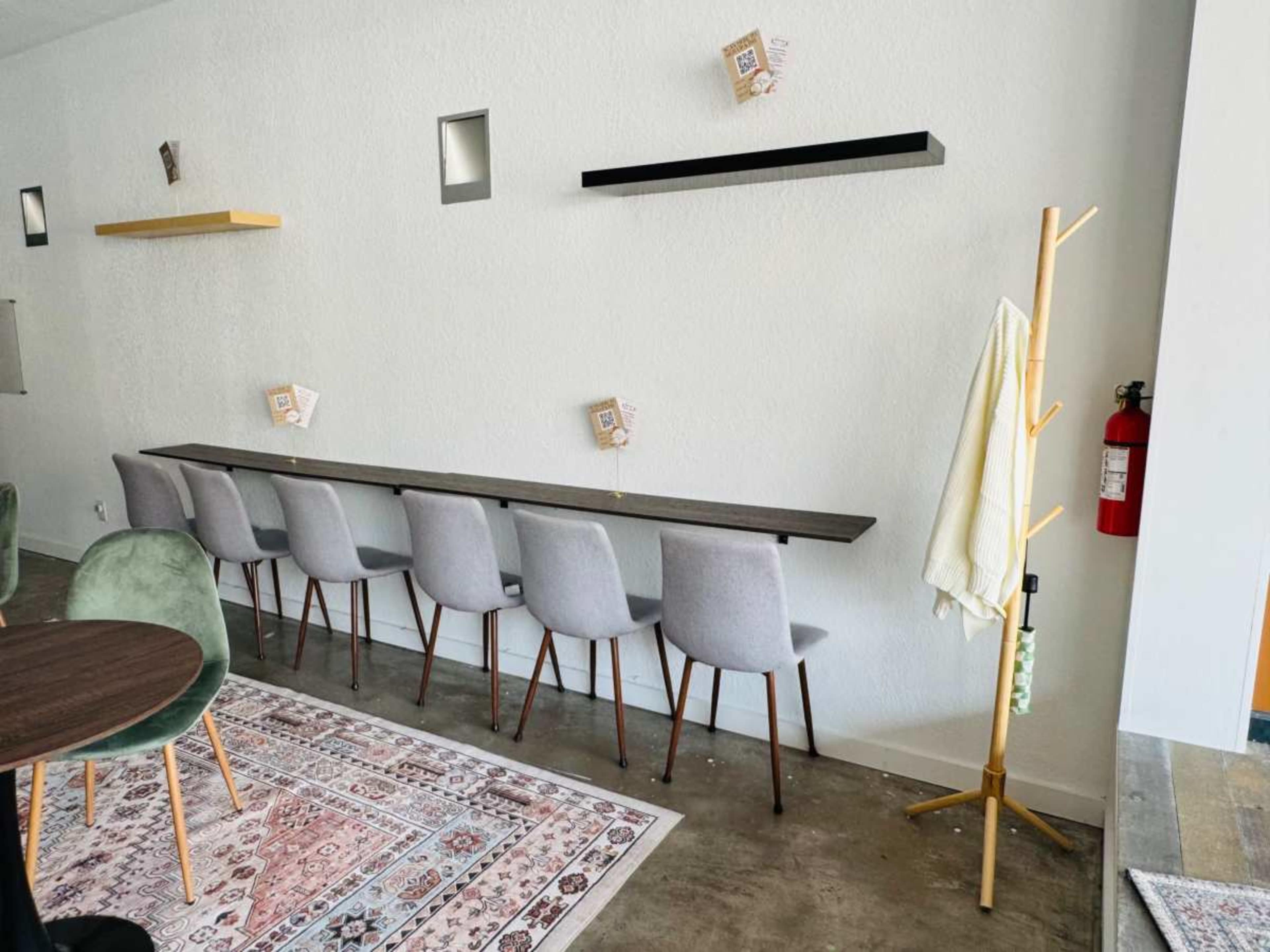 A minimalistic interior of a café featuring a long bar with several gray chairs, a wall-mounted shelf, and a wooden coat rack.