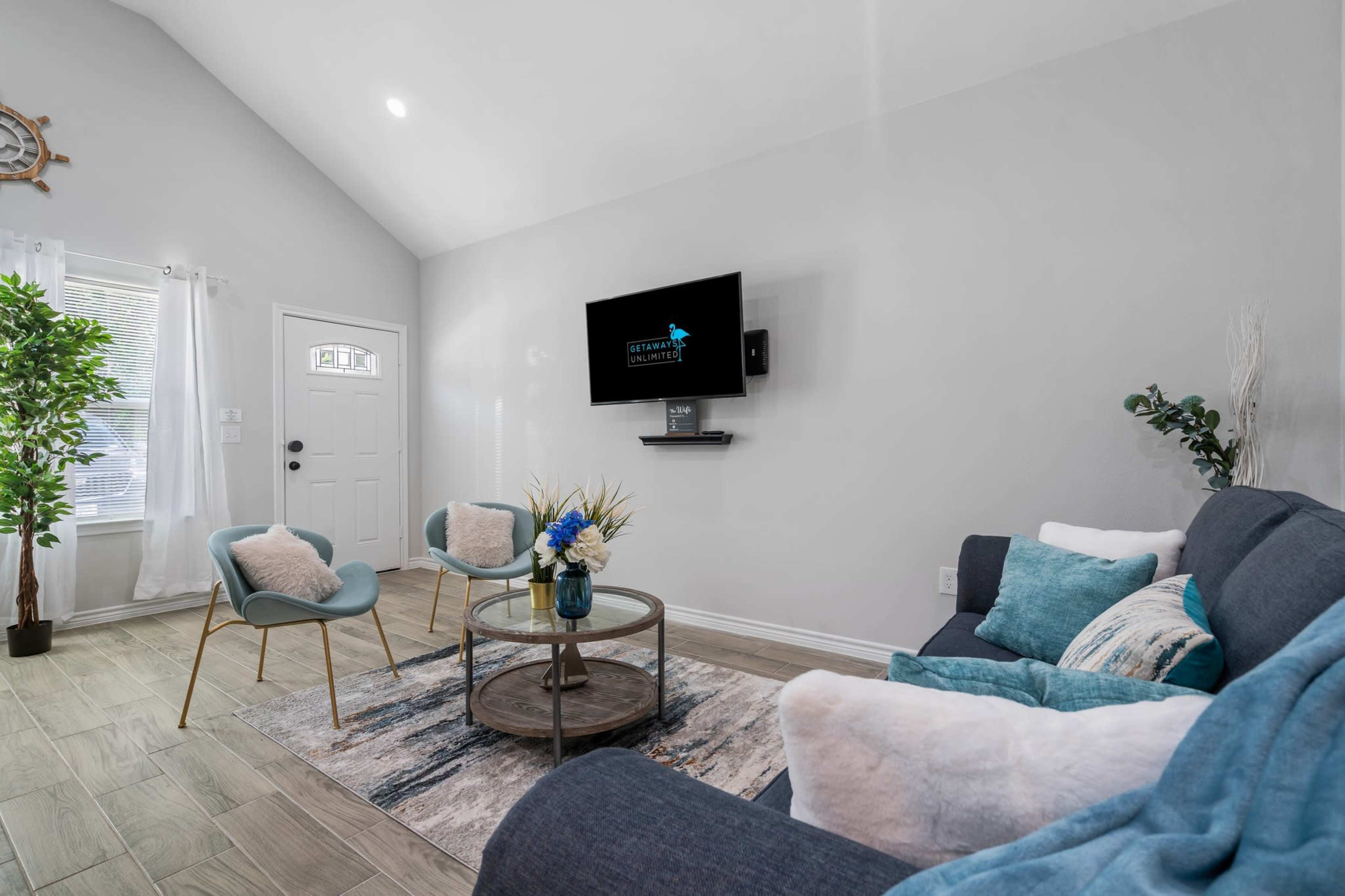 The living room features a blue sofa, two light blue chairs, a round coffee table with a vase of flowers, and a mounted television on a light gray wall.